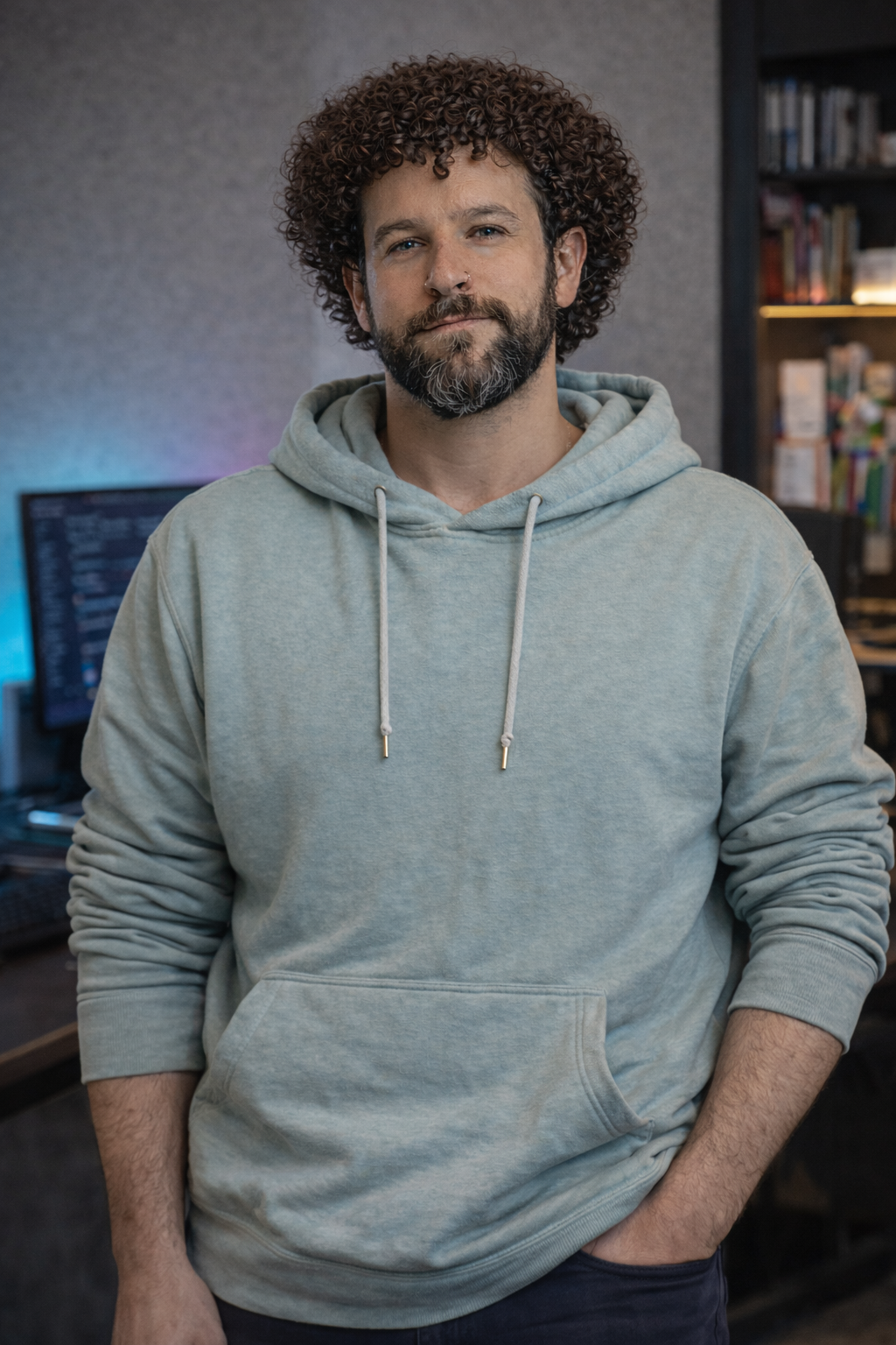 A man with curly brown hair and a beard, wearing a light gray hoodie, standing in a room with a blurred background of bookshelves and a computer monitor.