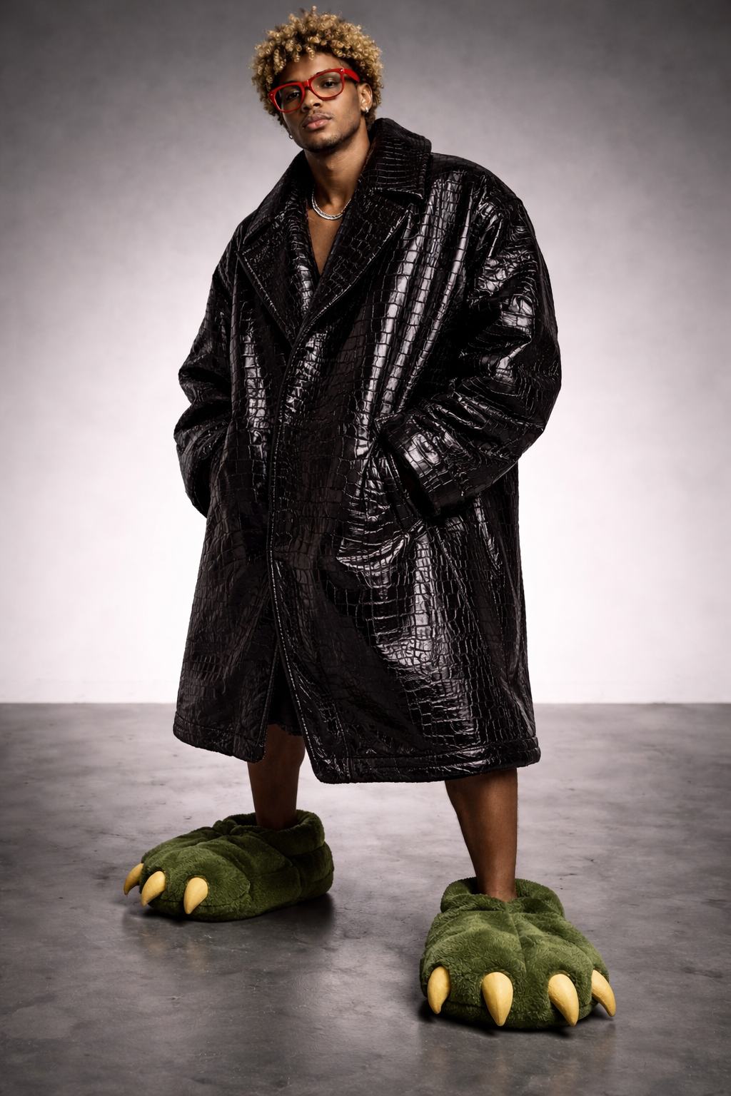 A person standing indoors wearing a black crocodile-patterned coat and green monster slippers with claws, red glasses, and a silver necklace, against a plain, light gray background.