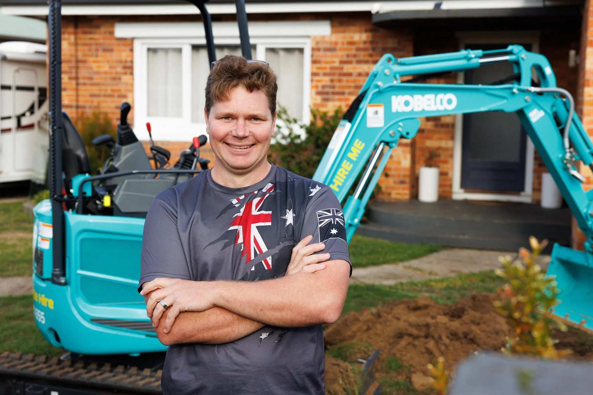 A smiling man with crossed arms standing in front of a blue mini excavator outside a brick house.