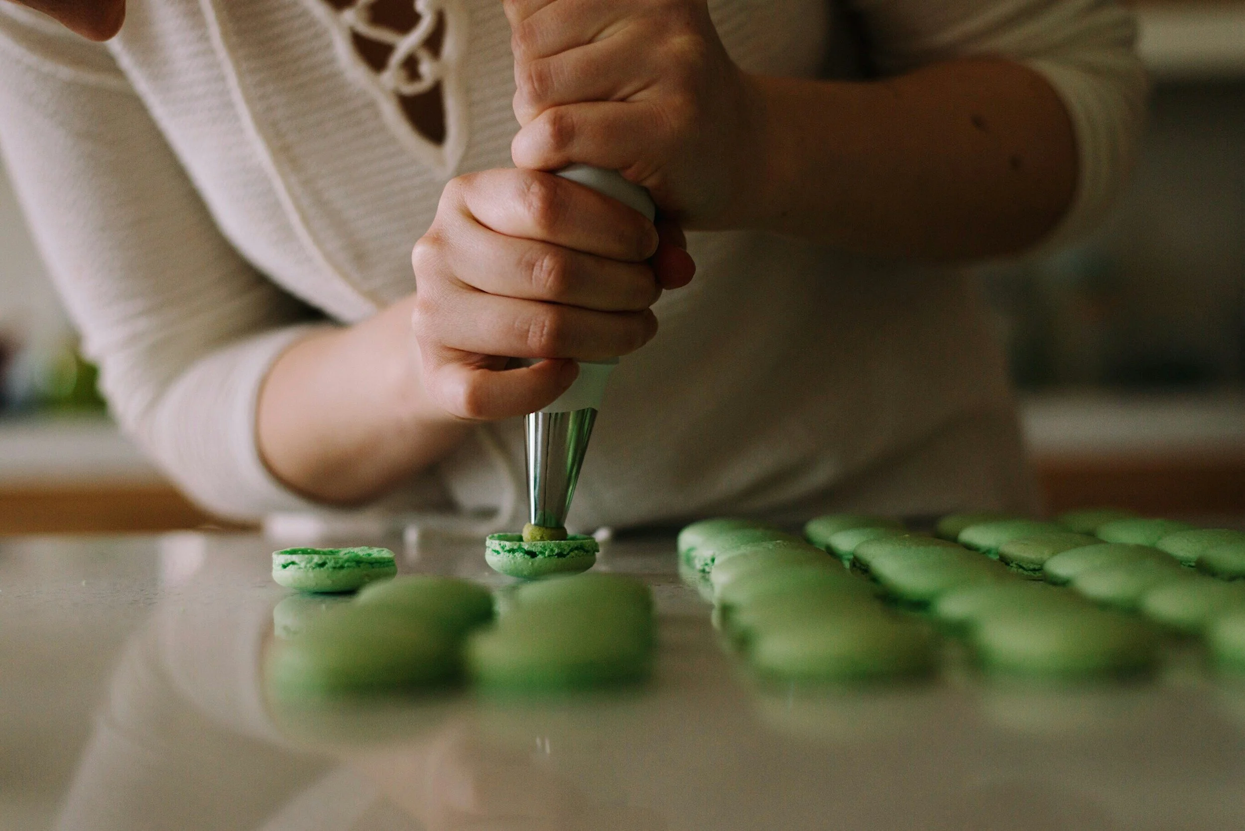 Close-up of a person decorating green macarons with a piping bag on a table.
