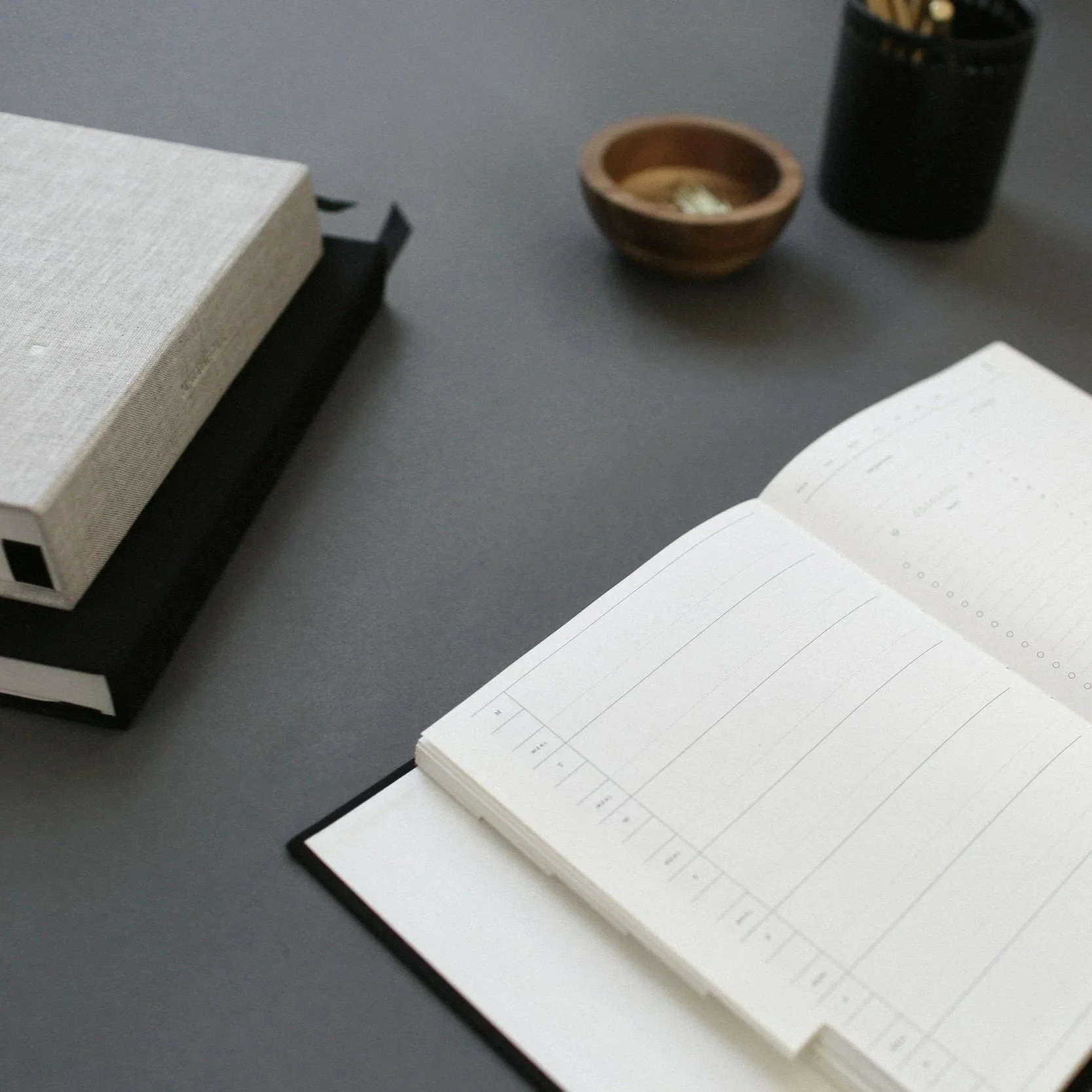 Open notebook with blank pages, a box, a small wooden bowl, and a black container on a gray surface.