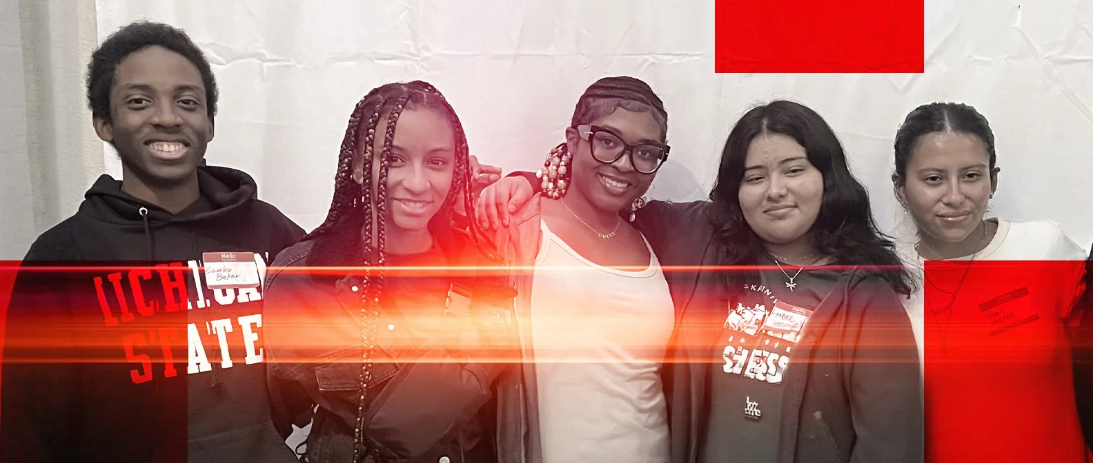 Group of five diverse young women standing together indoors, smiling at the camera, with a white backdrop and red banner above.