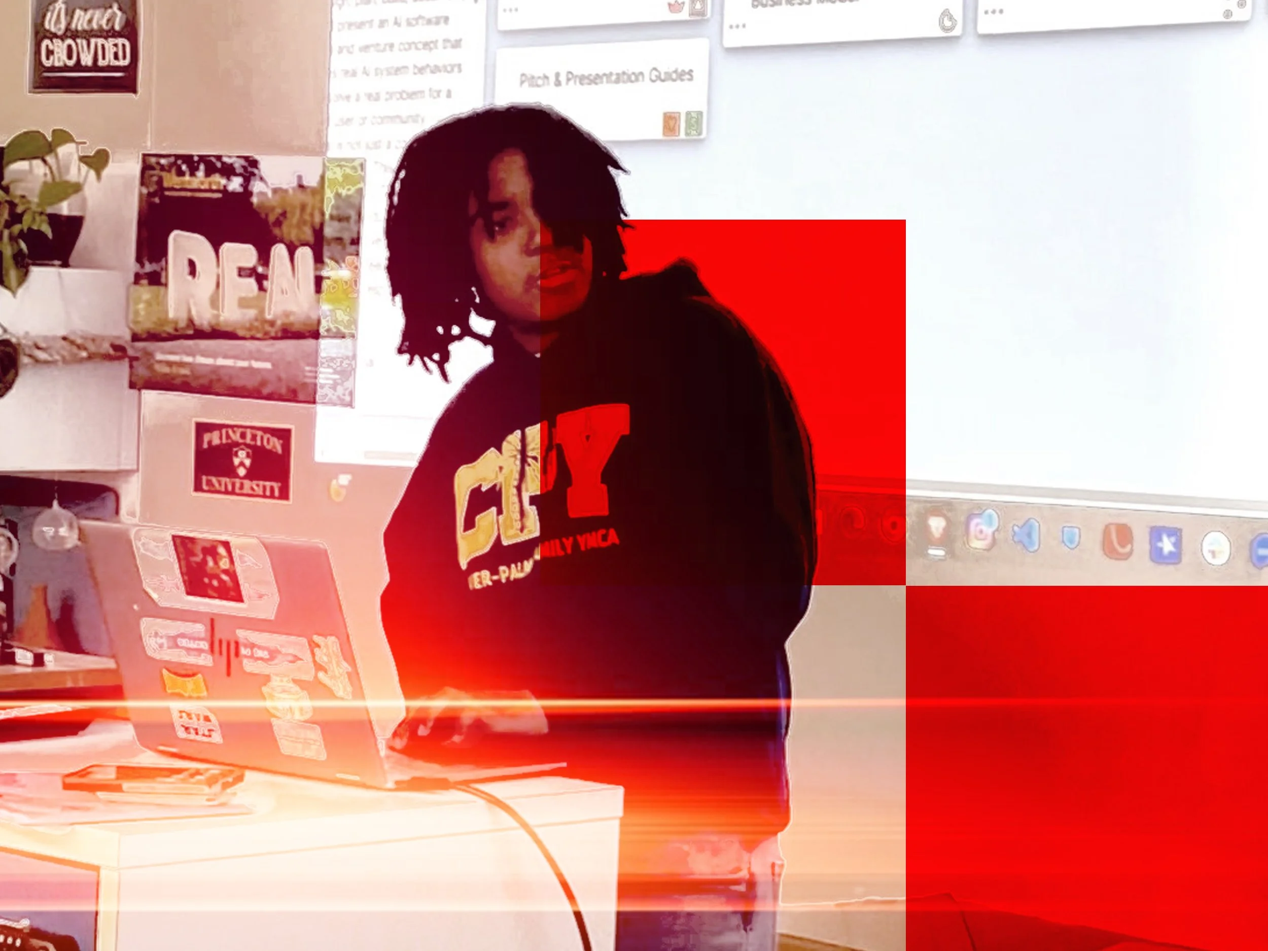 A person with dreadlocks standing in front of a whiteboard in a classroom or office setting, with various posters and signs on the wall, including a Princeton University logo and a sign that says 'Reach'.