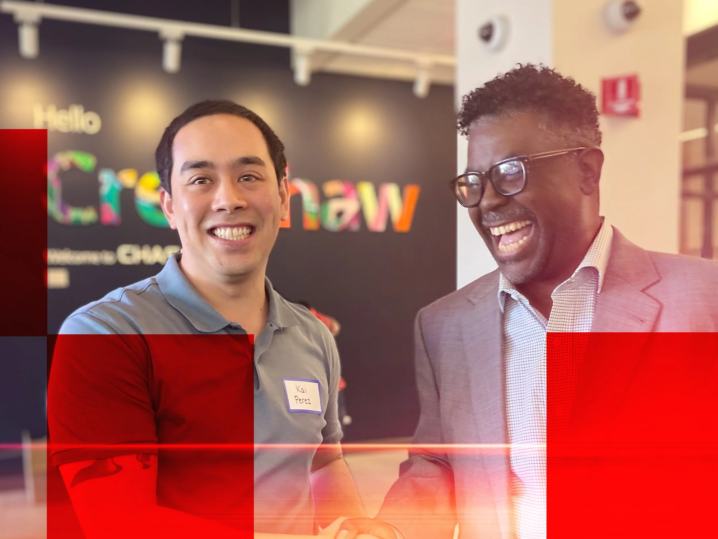 Two men, one of Asian descent and one of African descent, are smiling and engaging in conversation at an indoor event. The man on the left is wearing a gray polo shirt with a name tag, and the man on the right, who is laughing, is wearing glasses, a pink checkered shirt, and a brown blazer.