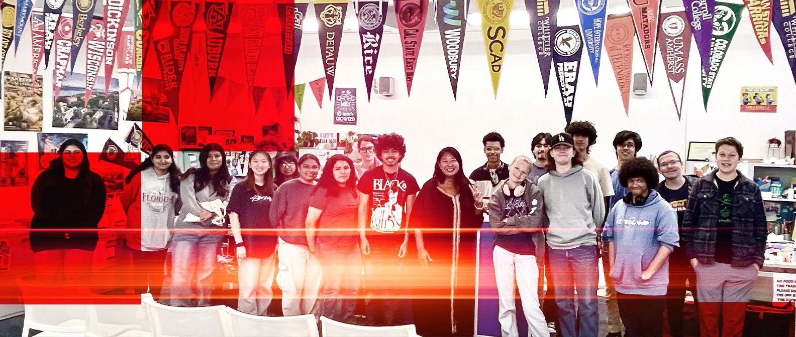 A group of diverse young people standing indoors in front of a wall decorated with university flags and posters, smiling at the camera.