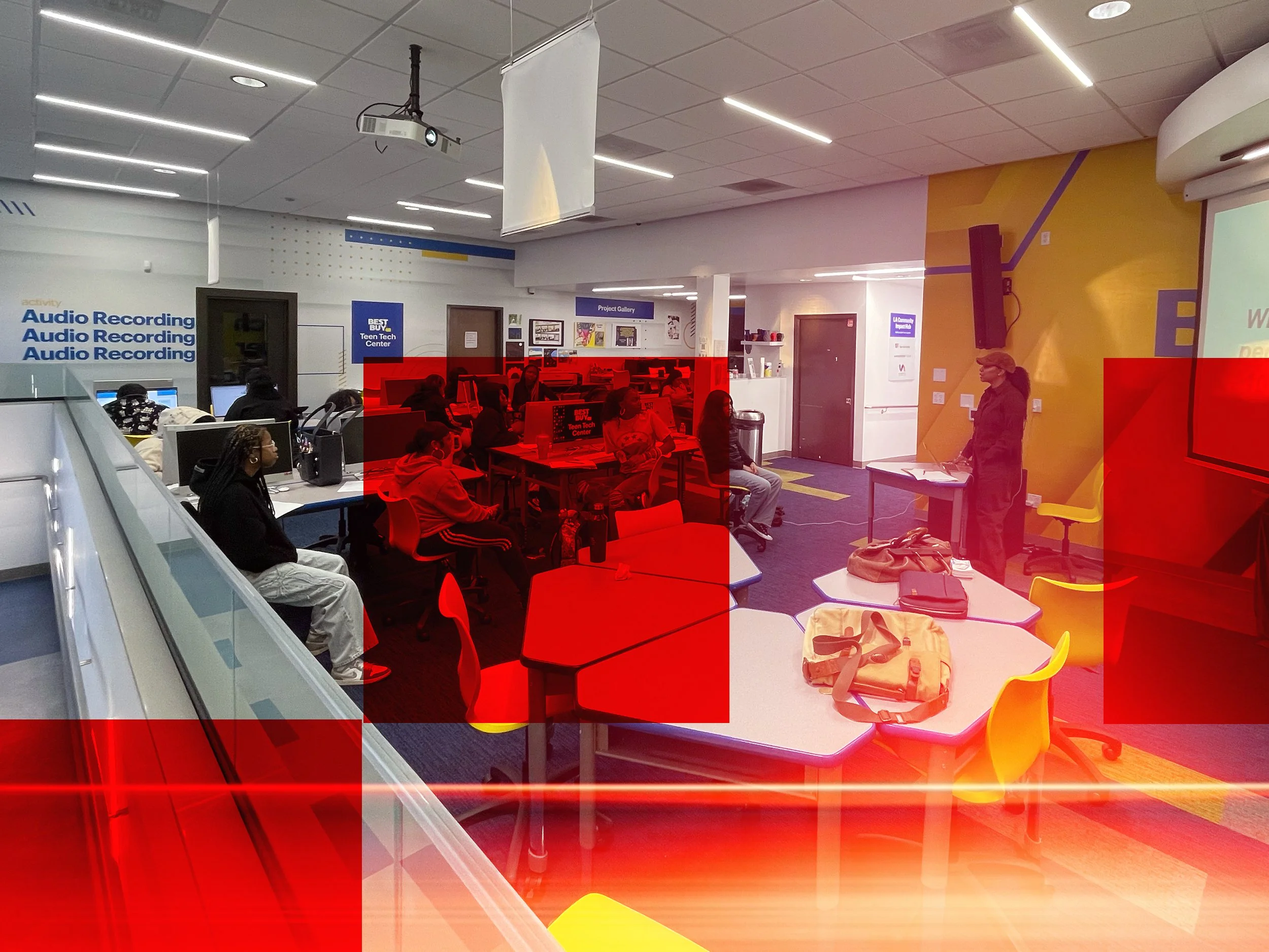 A computer lab or classroom with students seated at desks, some using computers, and a woman standing in front of the room near a projector screen. There are colorful chairs, backpacks, and educational posters on the walls.
