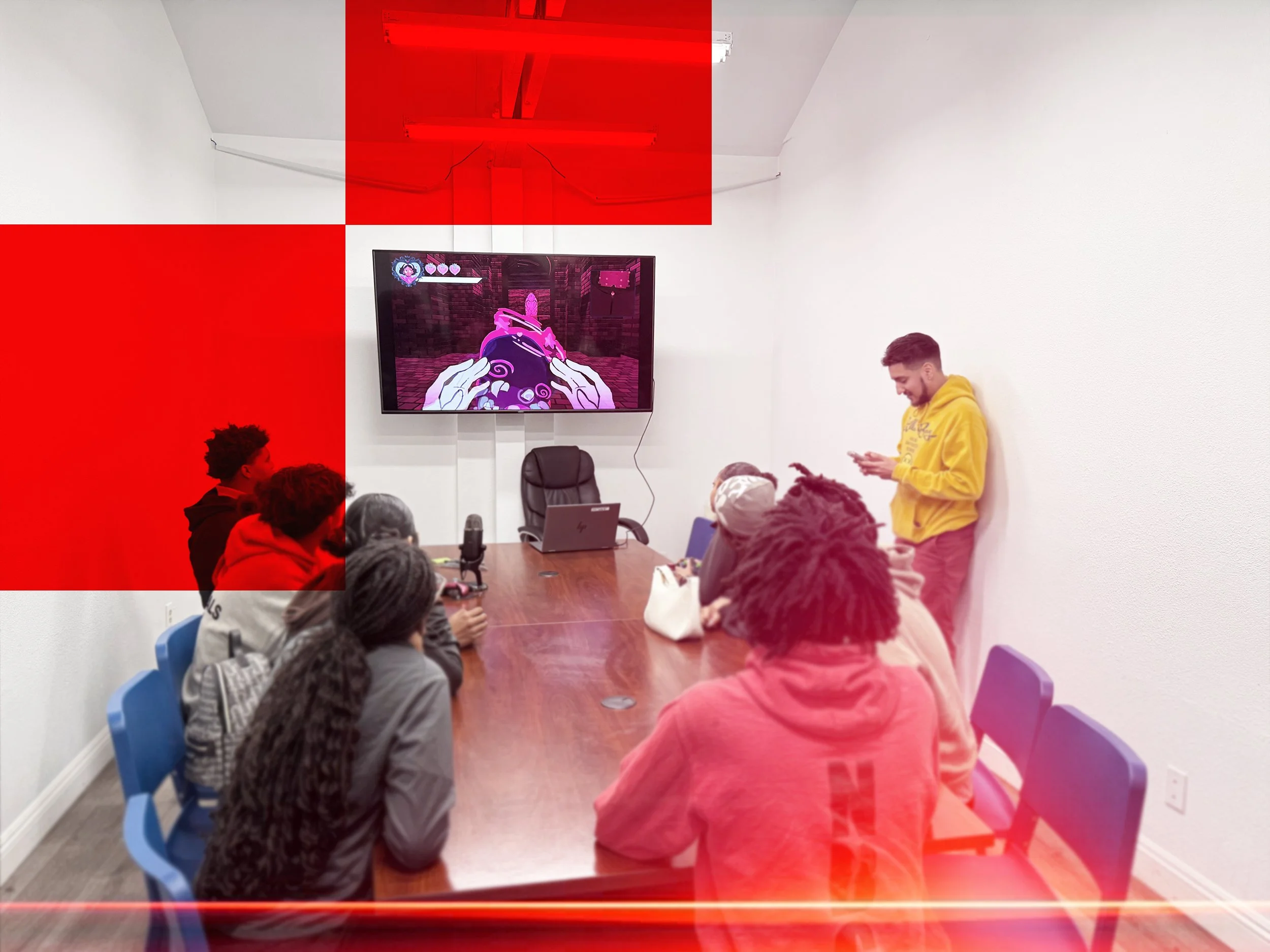 A group of people sitting around a conference table in a room with white walls. A man in a yellow hoodie is standing against the wall, using his phone. On the wall, there is a large screen display showing a video game with pink and purple visuals. Some red overlay sections are obscuring parts of the image.