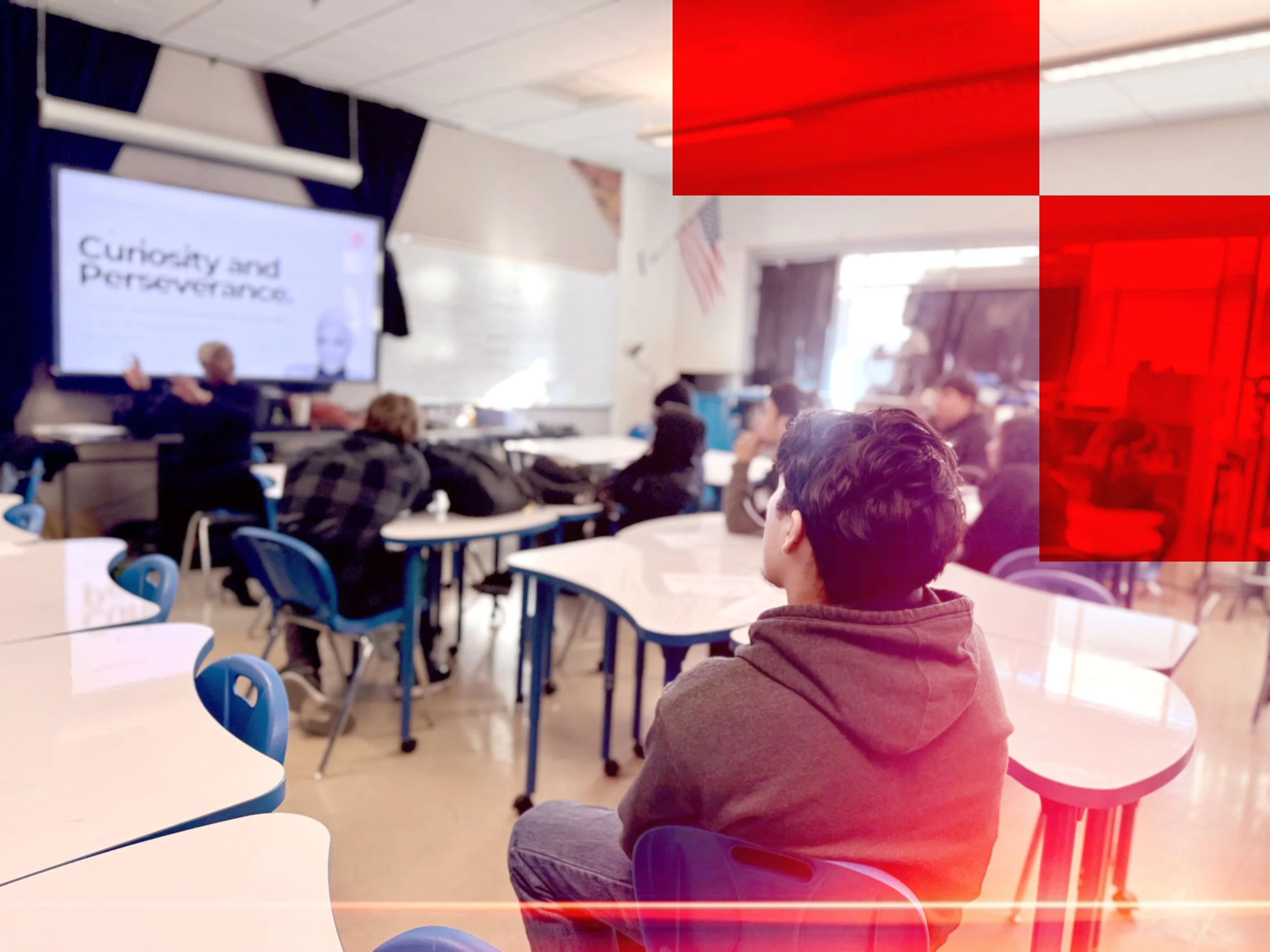 Students seated in a classroom or cafeteria, with a presenter or teacher at the front, and a screen displaying the words 'Curiosity and Perseverance.'