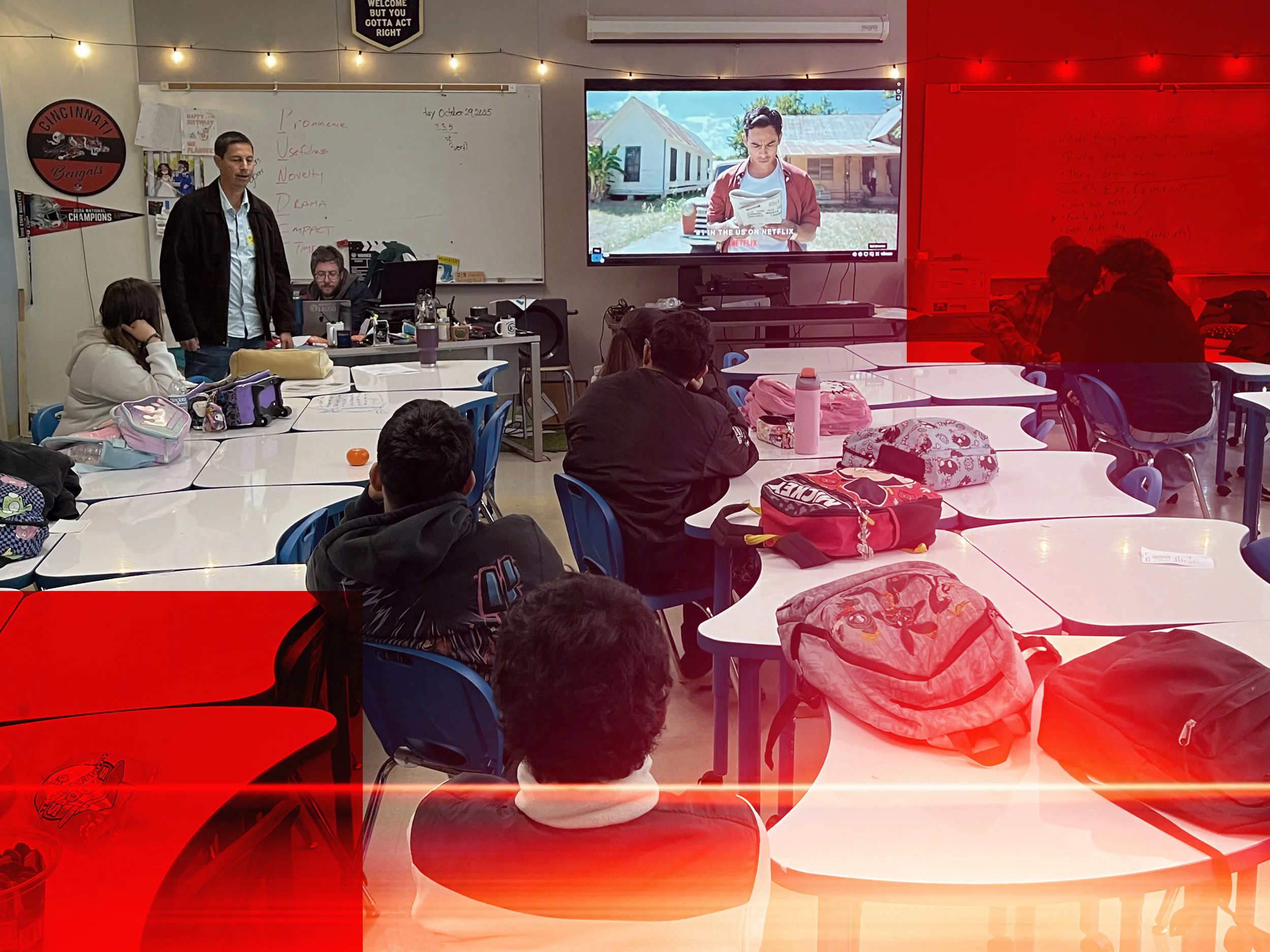 A classroom with students sitting at tables, a teacher standing, and a large screen showing a man reading a document outside, with two students on the right near a whiteboard. There are backpacks and water bottles on the tables.