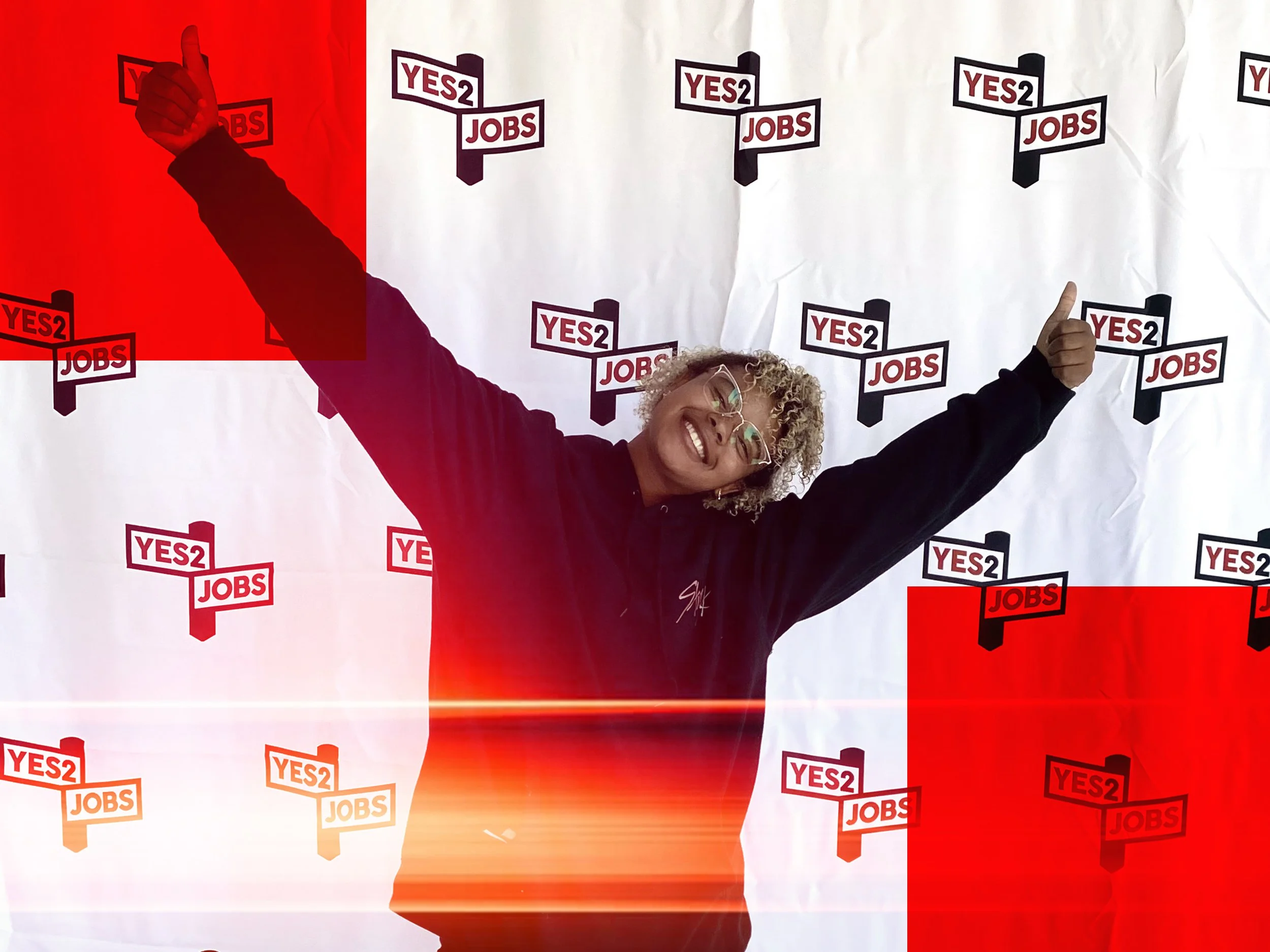 A woman with glasses smiling and posing with her arms outstretched in front of a background with multiple 'YES2 JOBS' logos. The background appears to be crumpled white paper, and there is a red overlay on some parts of the image.