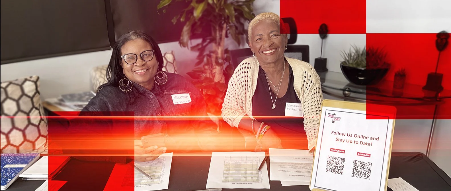 Two women smiling and sitting at a table with documents, tablet, and pens, in an indoor office or meeting space with plants and artwork in the background.