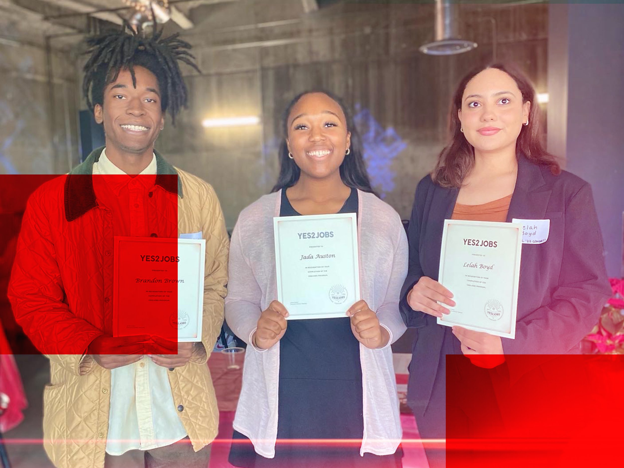 Three women standing side by side, each holding a certificate that reads "YES2JOBS" with their names: Brandon Brown, Jada Auston, and Lelah Boyd. They are smiling and appear happy at an indoor event with a wooden background.