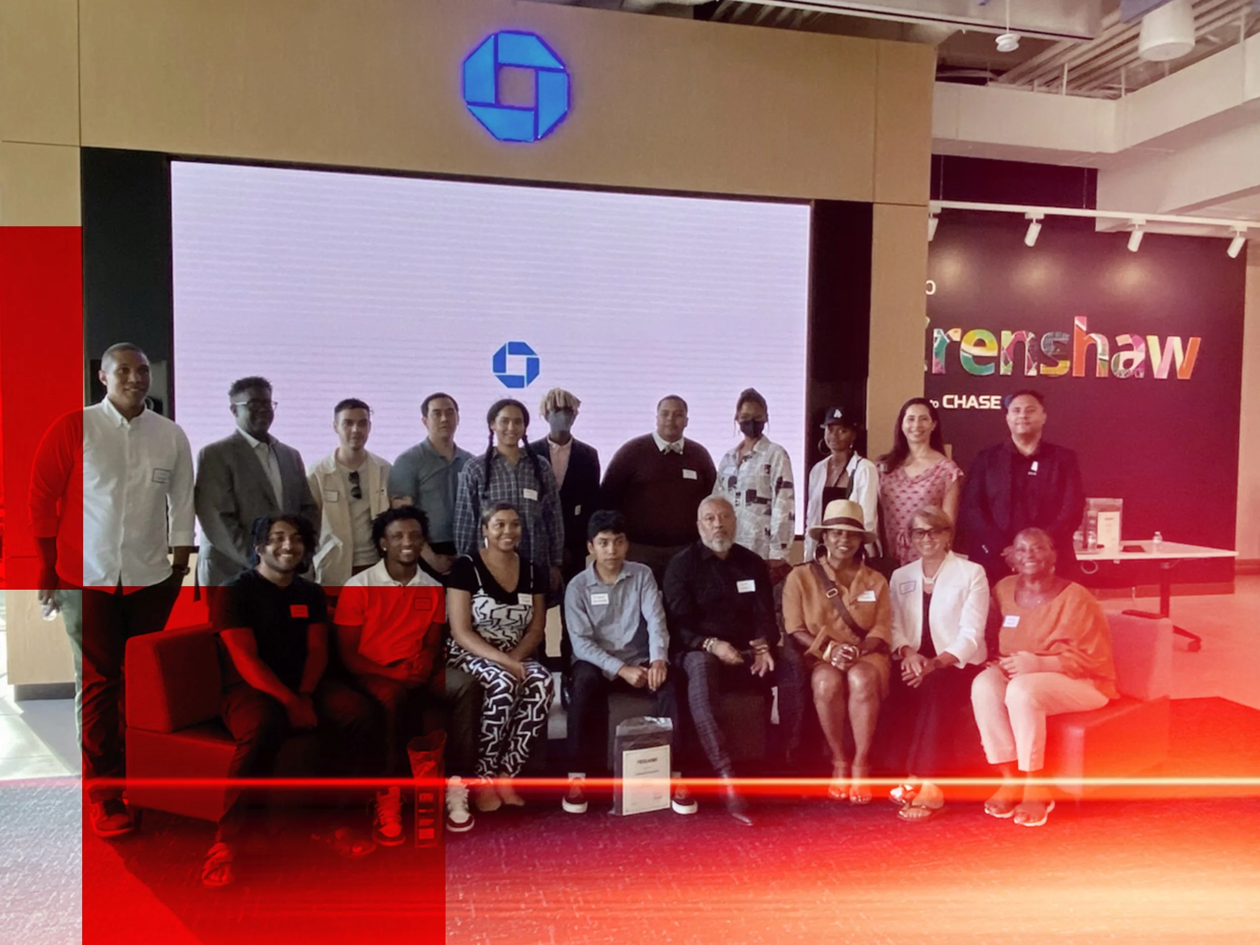 A group of diverse people standing and sitting in front of a large screen at an indoor event. Some individuals are smiling, wearing name tags, and casual or business casual attire. There is a Chase logo, a colorful sign that says 'Young Entrepreneurship', and people posing for a photo.