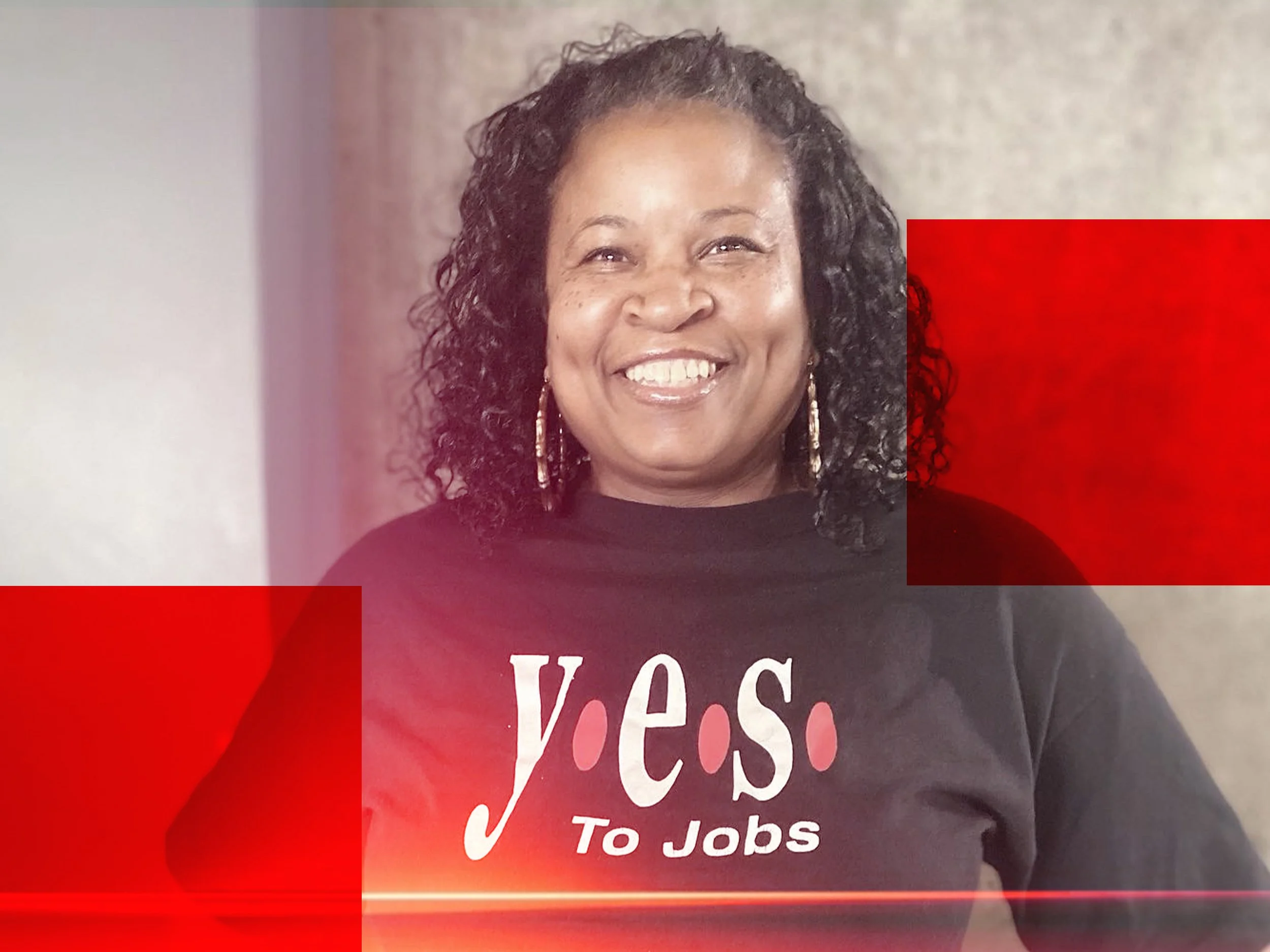 Close-up of a smiling woman with curly black hair wearing earrings and a black shirt with red and white text, against a background of beige and red.