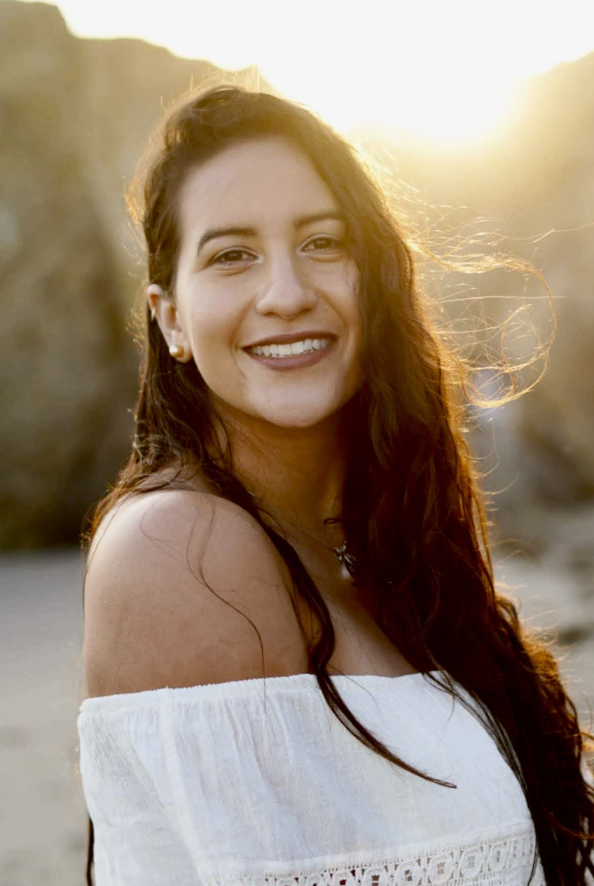 A woman smiling outdoors at sunset with long curly hair wearing a white off-shoulder top.