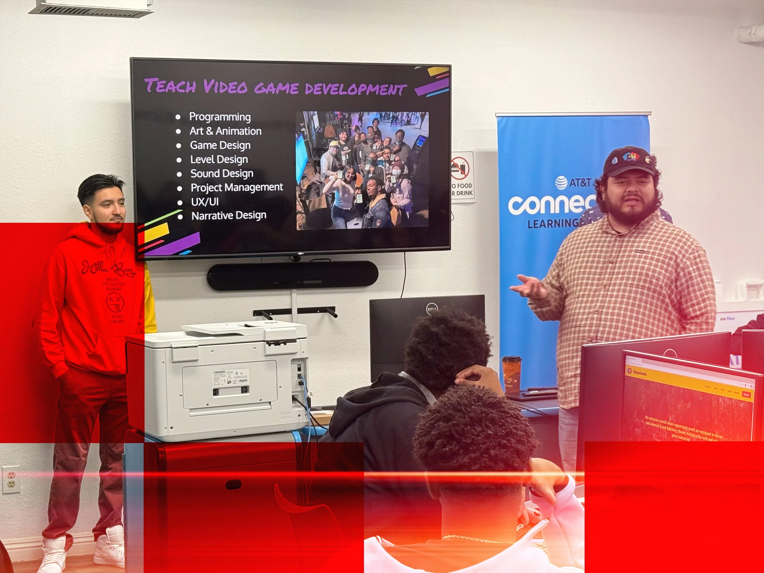 A man and a young man are standing in front of a presentation slide about video game development. The slide lists skills such as programming, art & animation, game design, level design, sound design, project management, UX/UI, and narrative design. The man is speaking and gesturing, and there are people seated and watching the presentation.