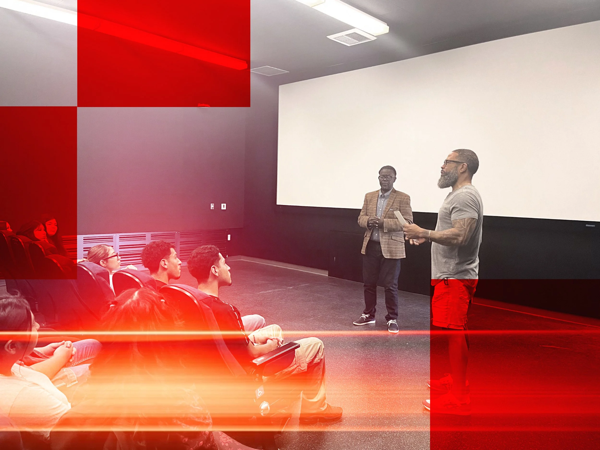 Two men are standing and speaking to an audience in a room with white and purple walls and a large blank screen behind them. The audience, consisting of multiple people, is seated and listening.