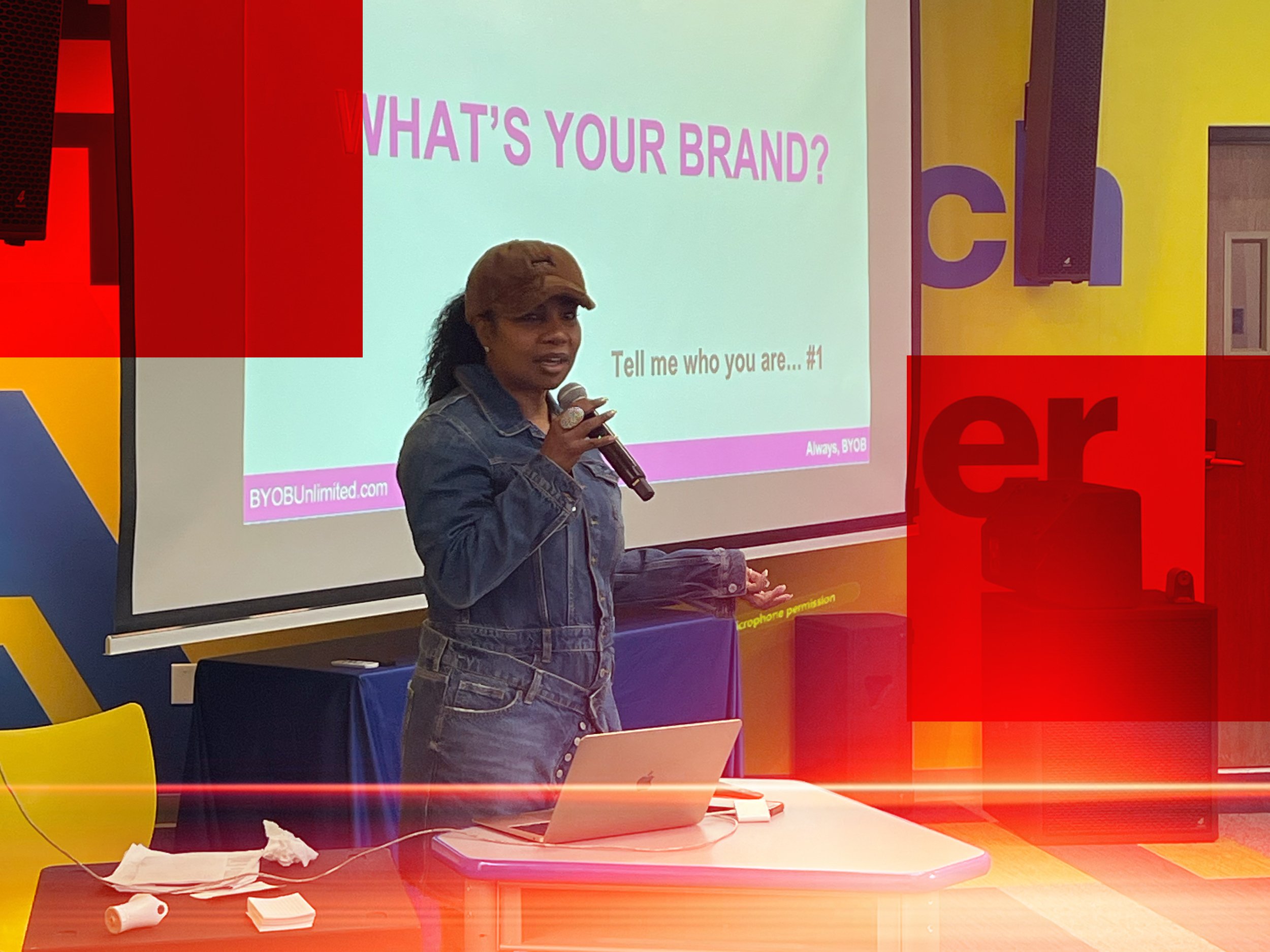 A woman with a denim jacket and a brown cap holding a microphone is standing in front of a screen that displays the text 'WHAT'S YOUR BRAND?'. There is a table with a laptop, tissue papers, and a pink object in the foreground. The background has colorful walls with large letter shapes.