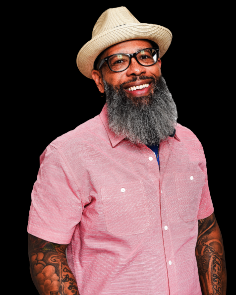 A smiling man with glasses, a beard, and tattoos, wearing a light-colored hat and pink short-sleeved shirt, against a black background.