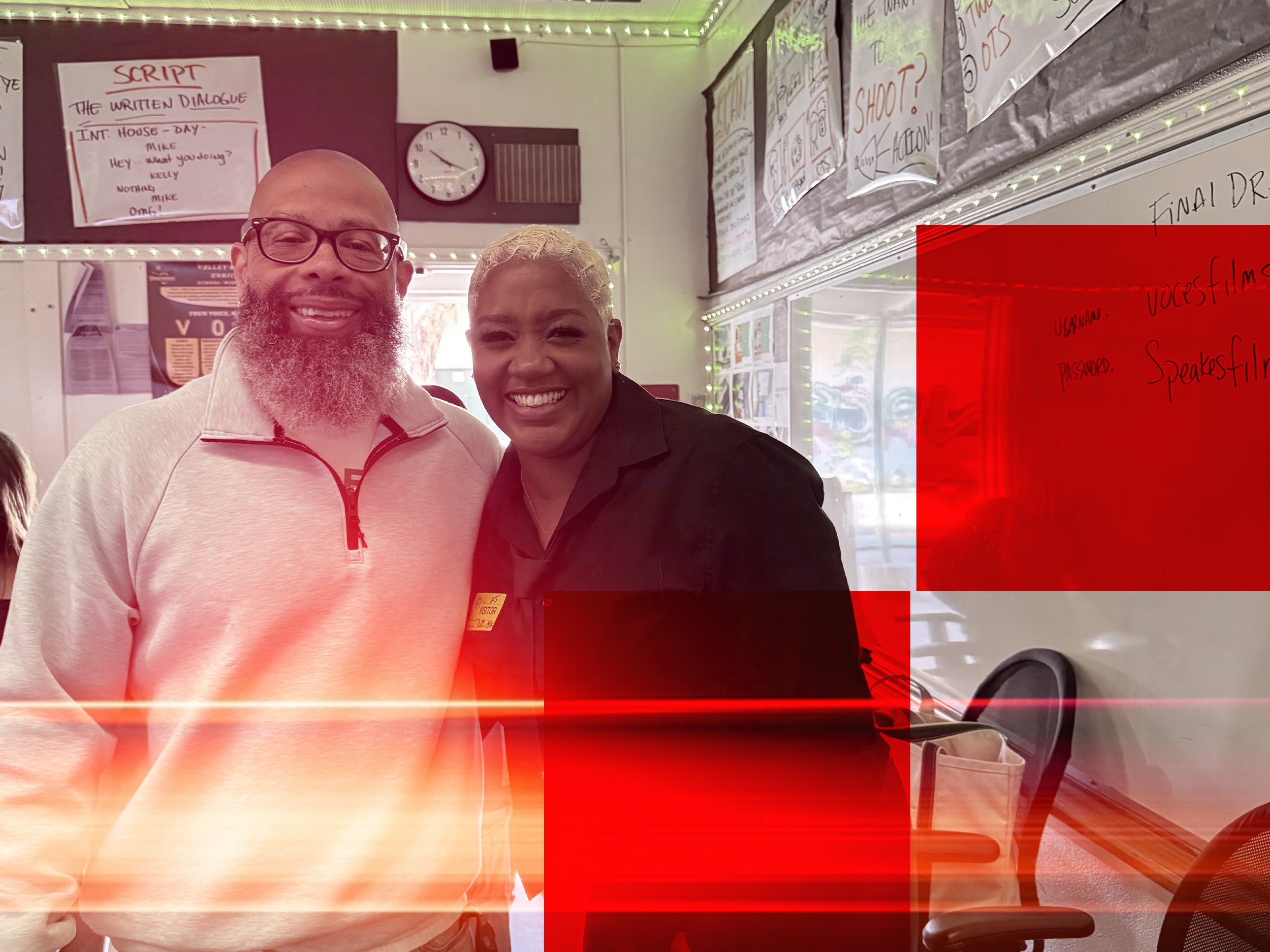 Two smiling people standing indoors near a wall with handwritten notes and posters, with a red overlay and light effects in the photo.