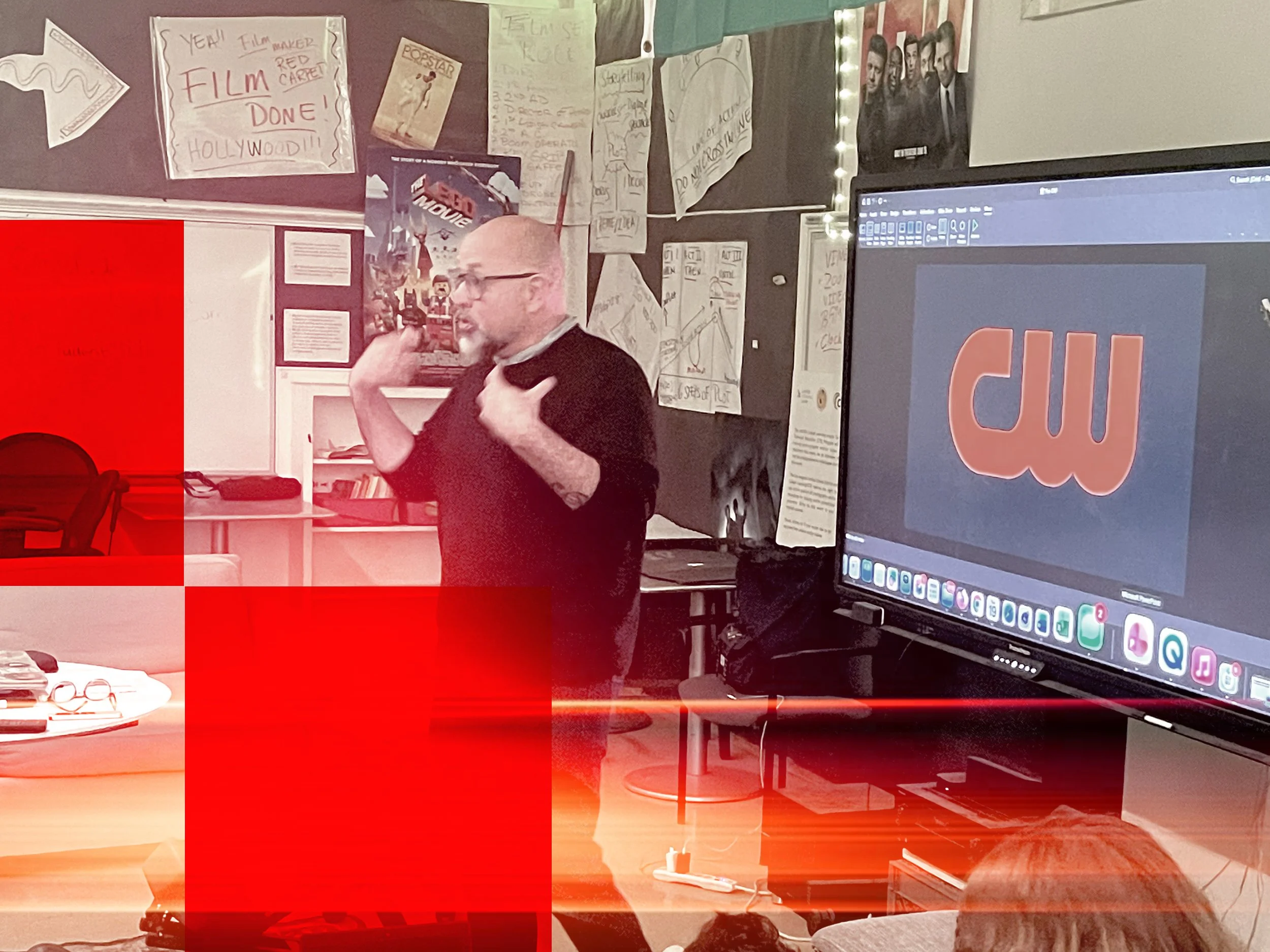 Person with glasses and a beard, wearing glasses and a dark shirt, standing in a classroom or presentation room, gesturing with his hand near his collar. The room has various posters and handwritten charts on the walls, a large monitor displaying a logo or design, and desks with scattered items. There are chairs and a computer on the table, indicating a tech or educational environment.