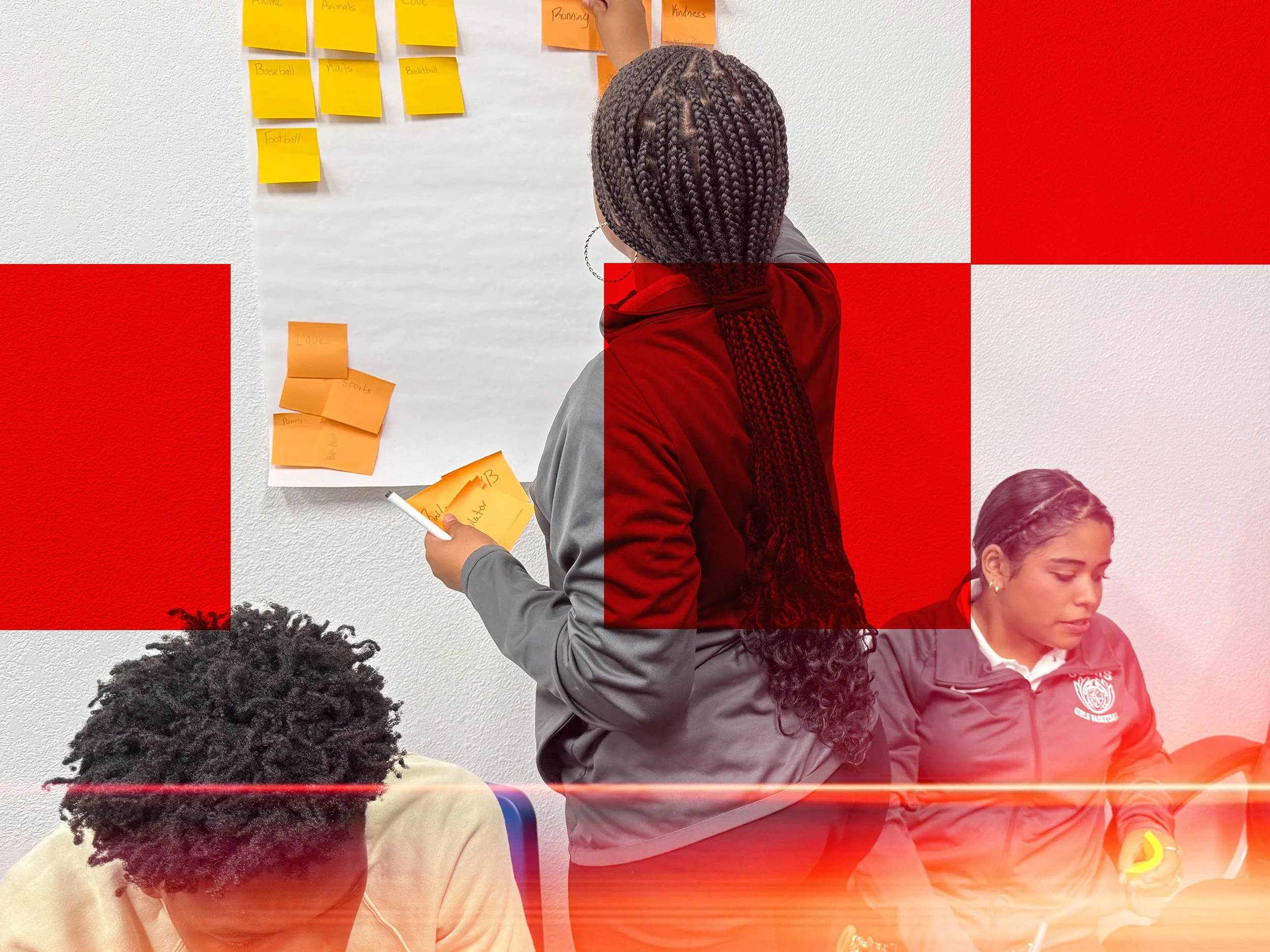 A person with braided hair writing on a whiteboard with sticky notes, surrounded by students seated at a table, in a classroom setting.
