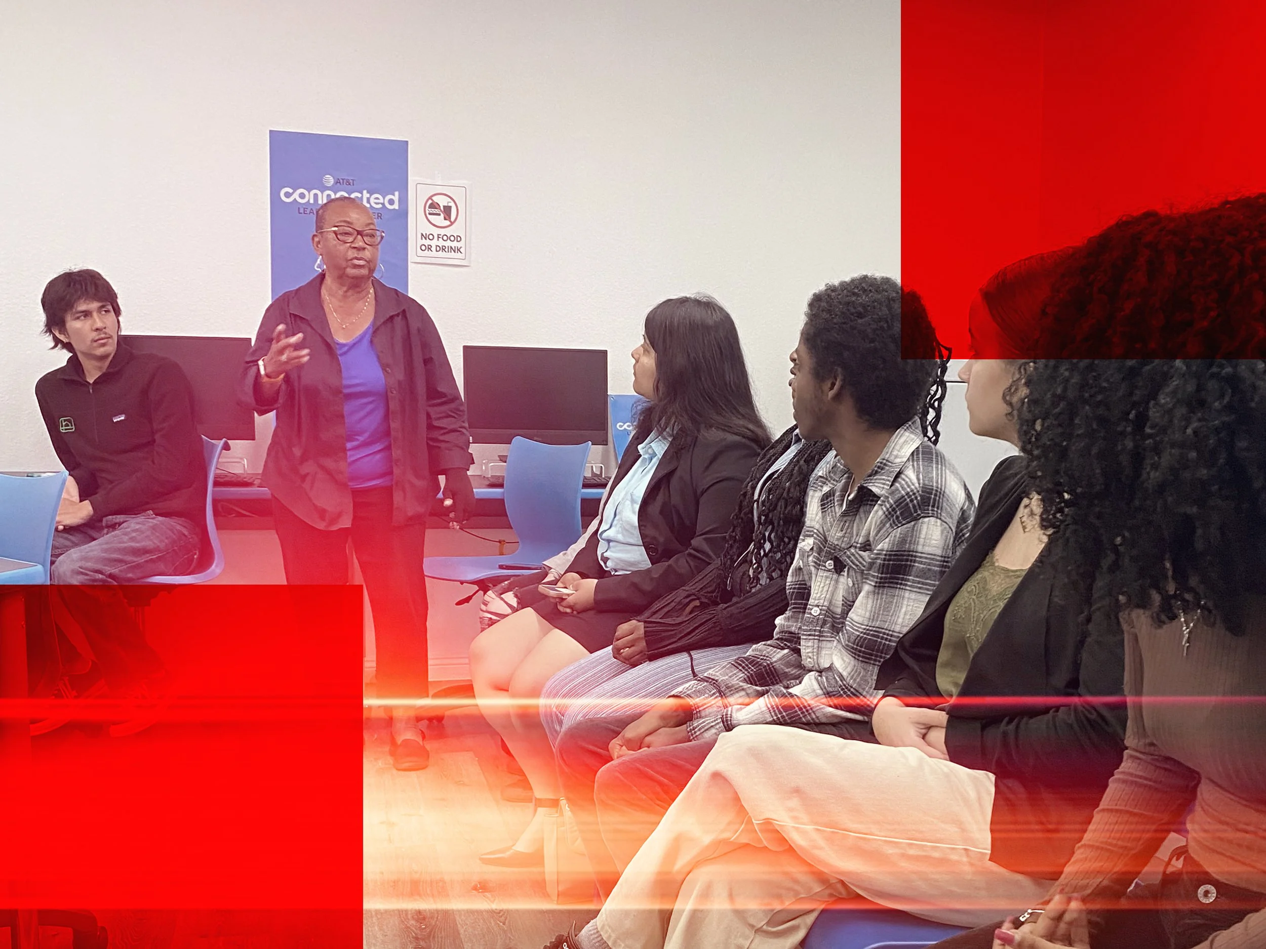 A woman speaking in front of a group of five seated people in a classroom or office setting. Behind her, there are two desktop computers and a poster that reads 'connected' and 'LEARN'. A sign on the wall indicates no food or drink allowed.