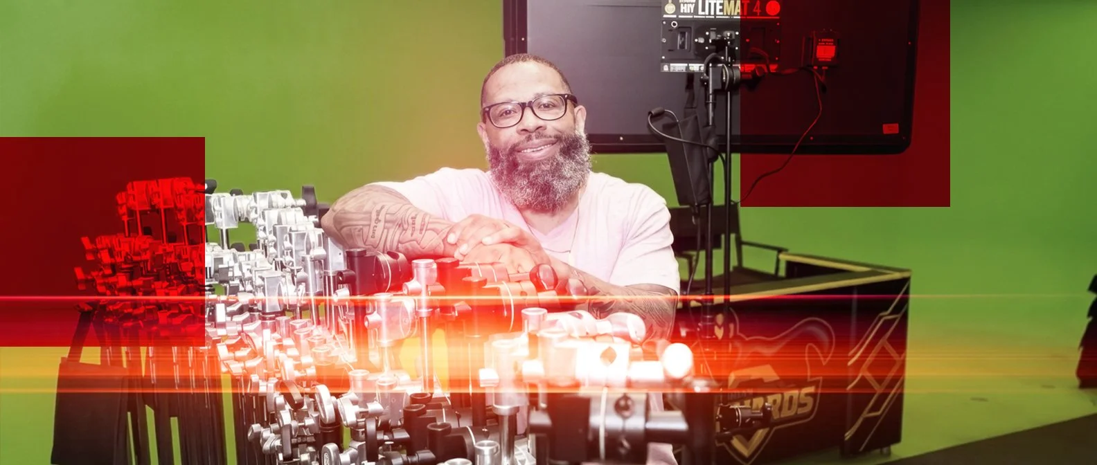 A man with glasses and a beard is smiling at a mix of robots and mechanical components on a table in a studio with green background and technical equipment.