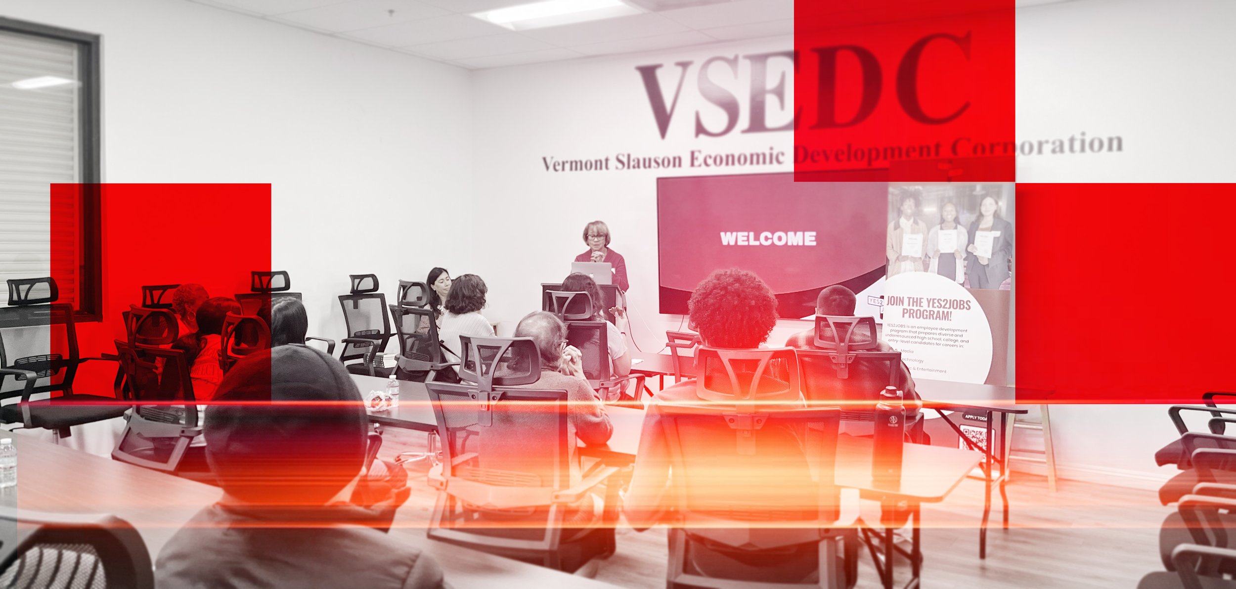 A woman speaking at a podium during a presentation in a conference room with attendees seated facing her. There is a large screen displaying 'WELCOME' and information about a job program.