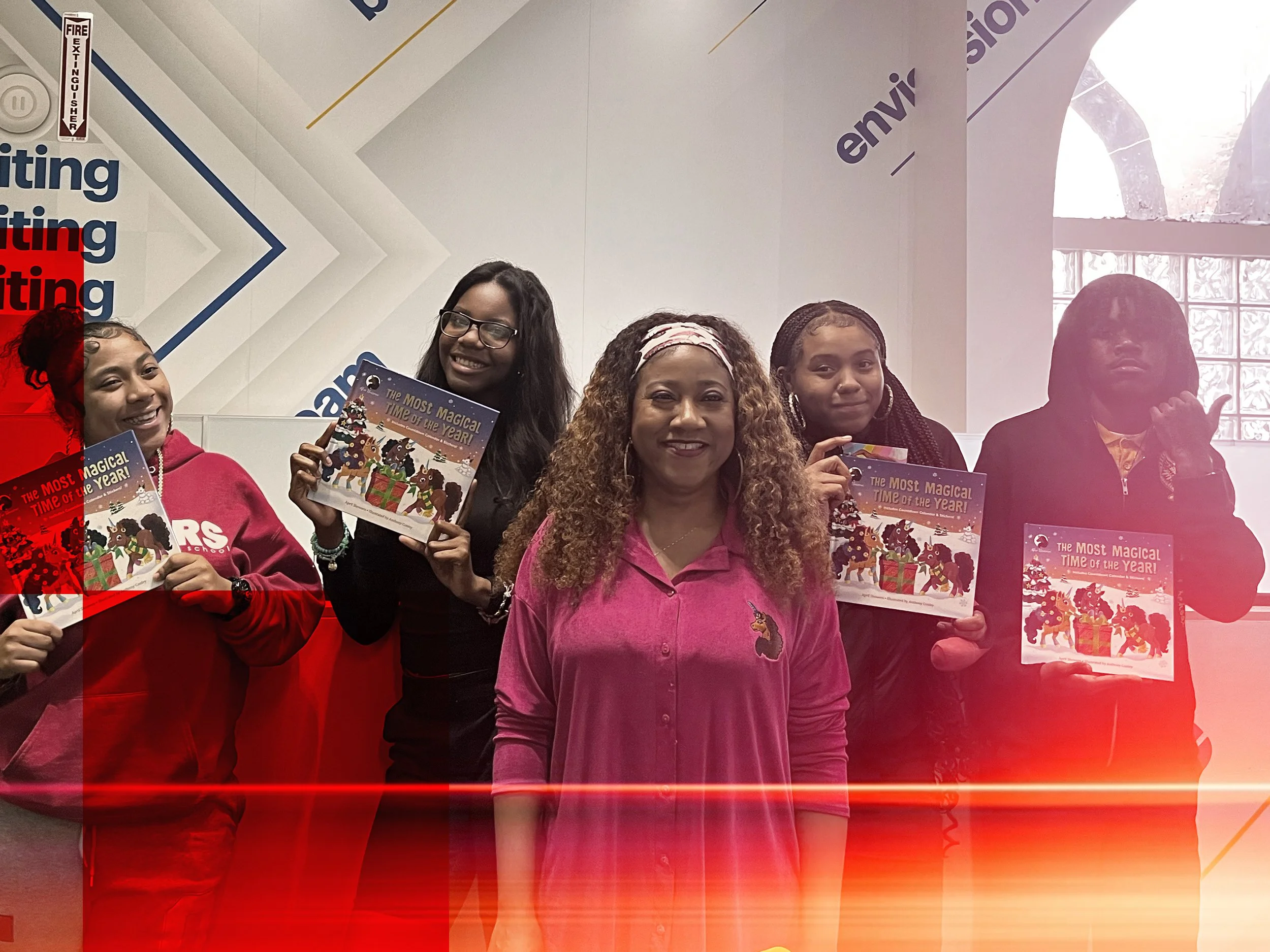 Group of five women posing indoors, holding colorful books titled "The Most Magical Time of the Year" with Christmas-themed illustrations. An indoor setting with graphic wall decor and large window in the background.