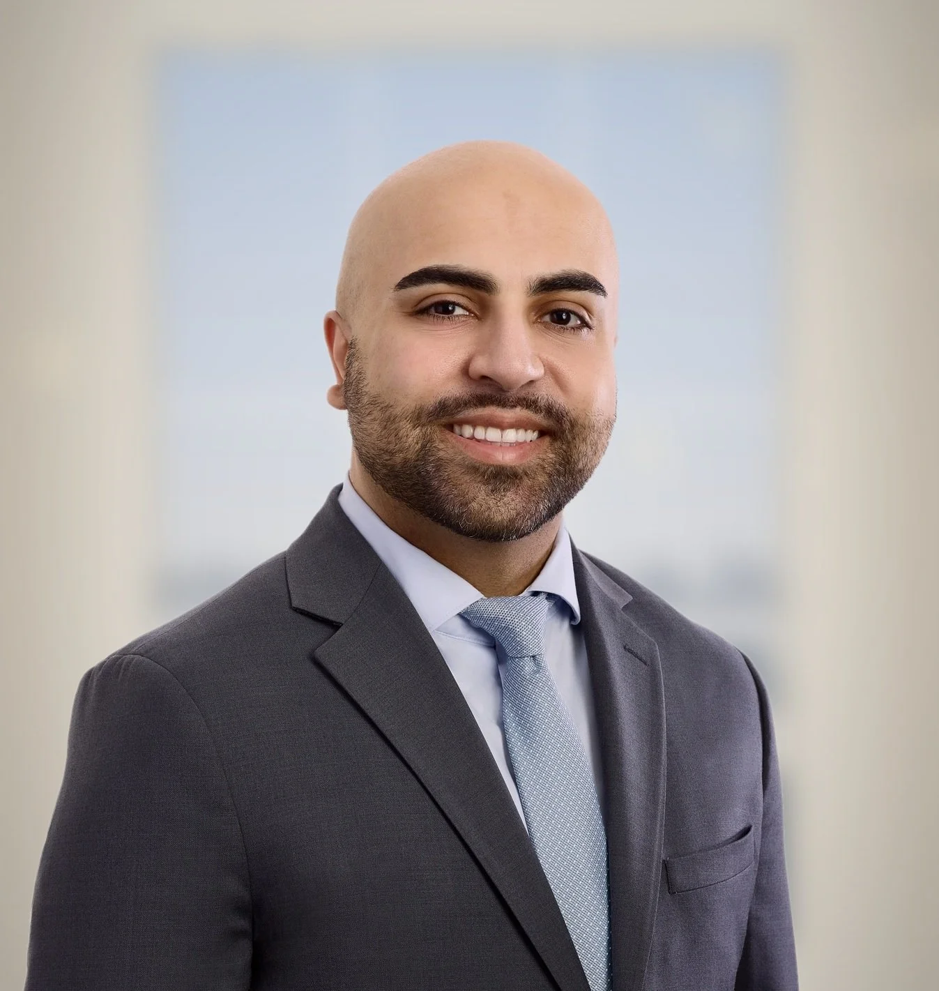Professional portrait of a bald man with a beard, wearing a gray suit, light blue shirt, and matching tie, smiling against a blurred light background.