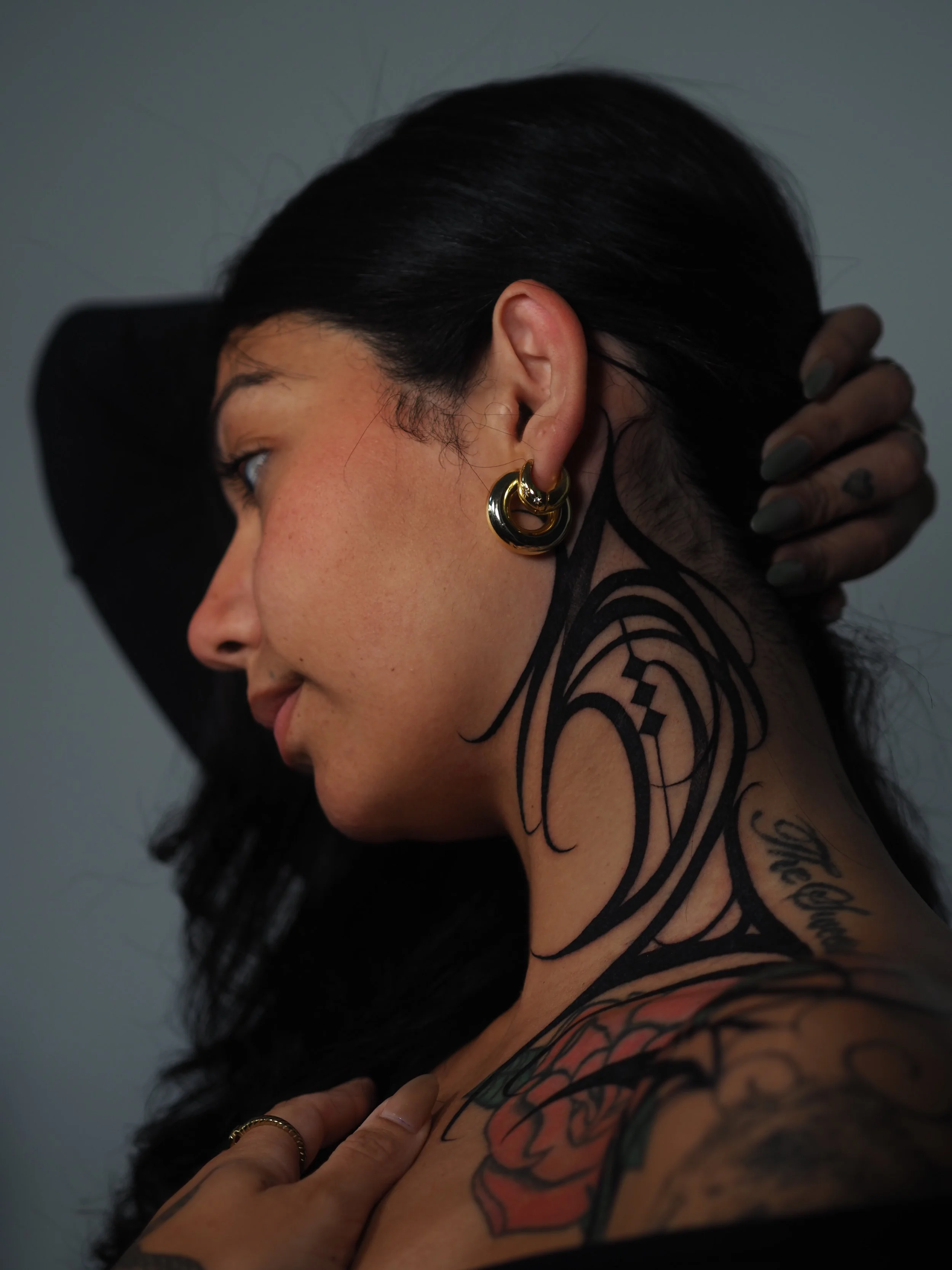 A woman with long black hair, wearing gold hoop earrings, running her hand through her hair and showing tattoos on her neck and shoulder.