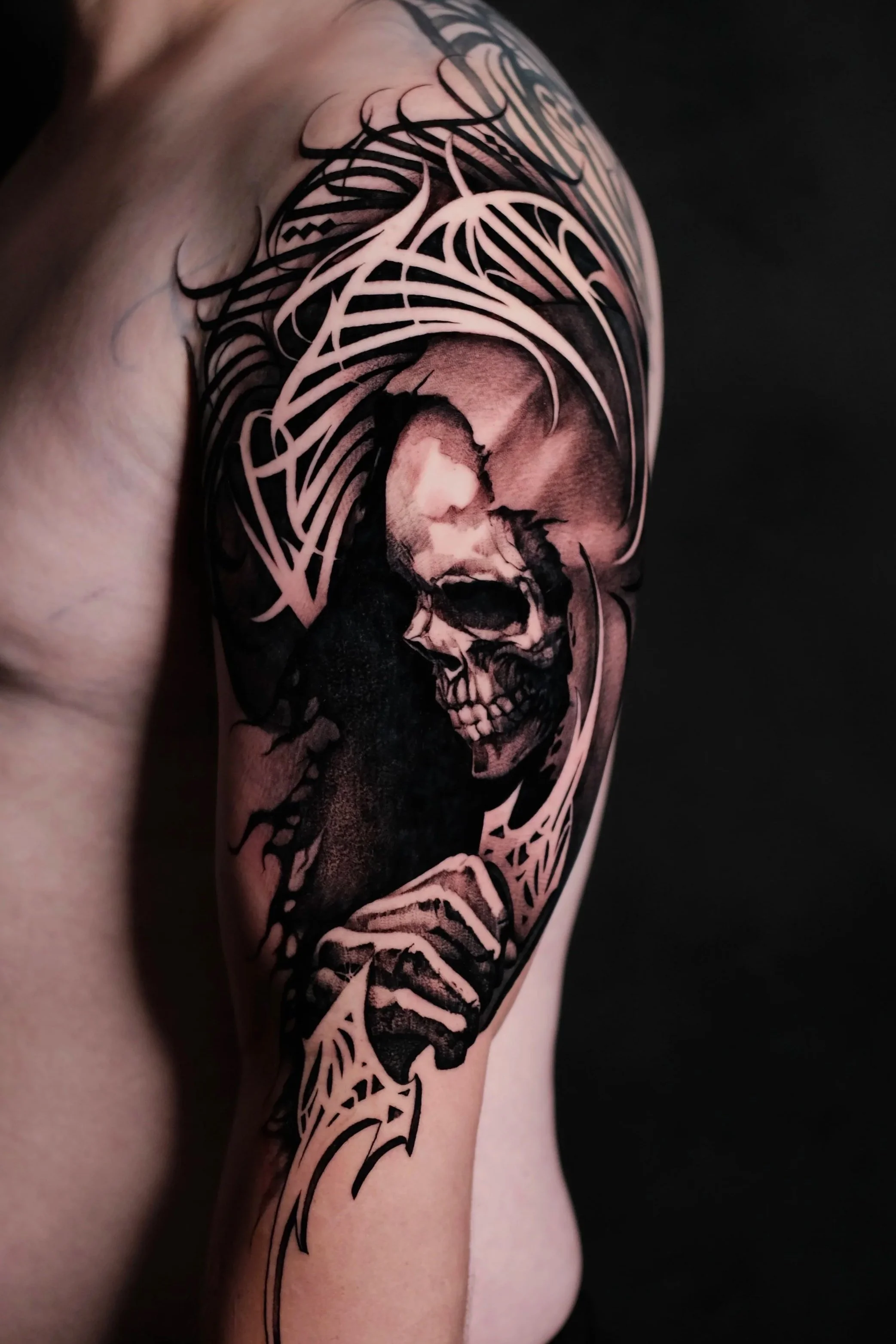 A detailed black and gray tattoo of a skull with abstract lines and shading on a person's upper arm and shoulder.