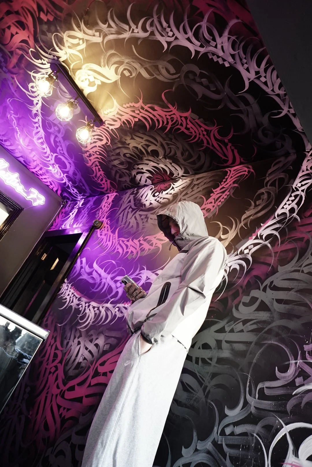 Person in a white hoodie and pants looking at a smartphone, standing against a wall with abstract pink and gray graffiti-style art and colorful neon lighting.