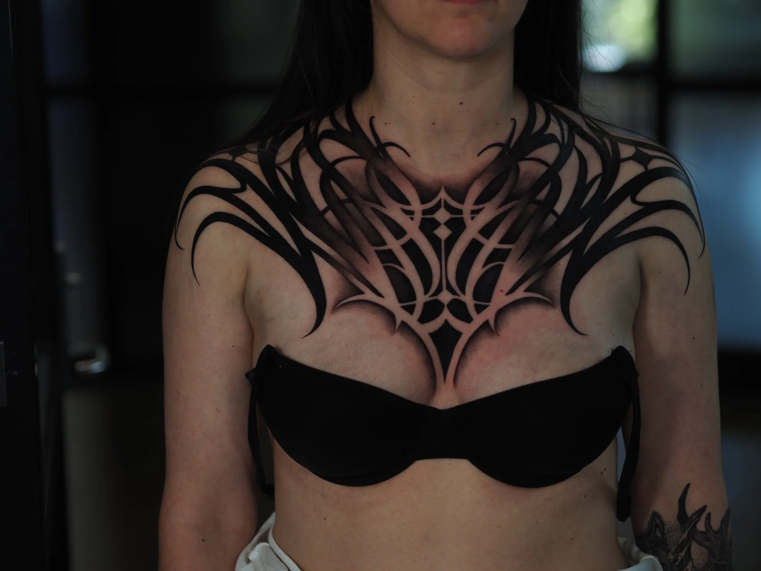 Close-up of a woman with black hair and large black tattoo resembling a spider web on her chest and shoulders, wearing a black top with brown straps.