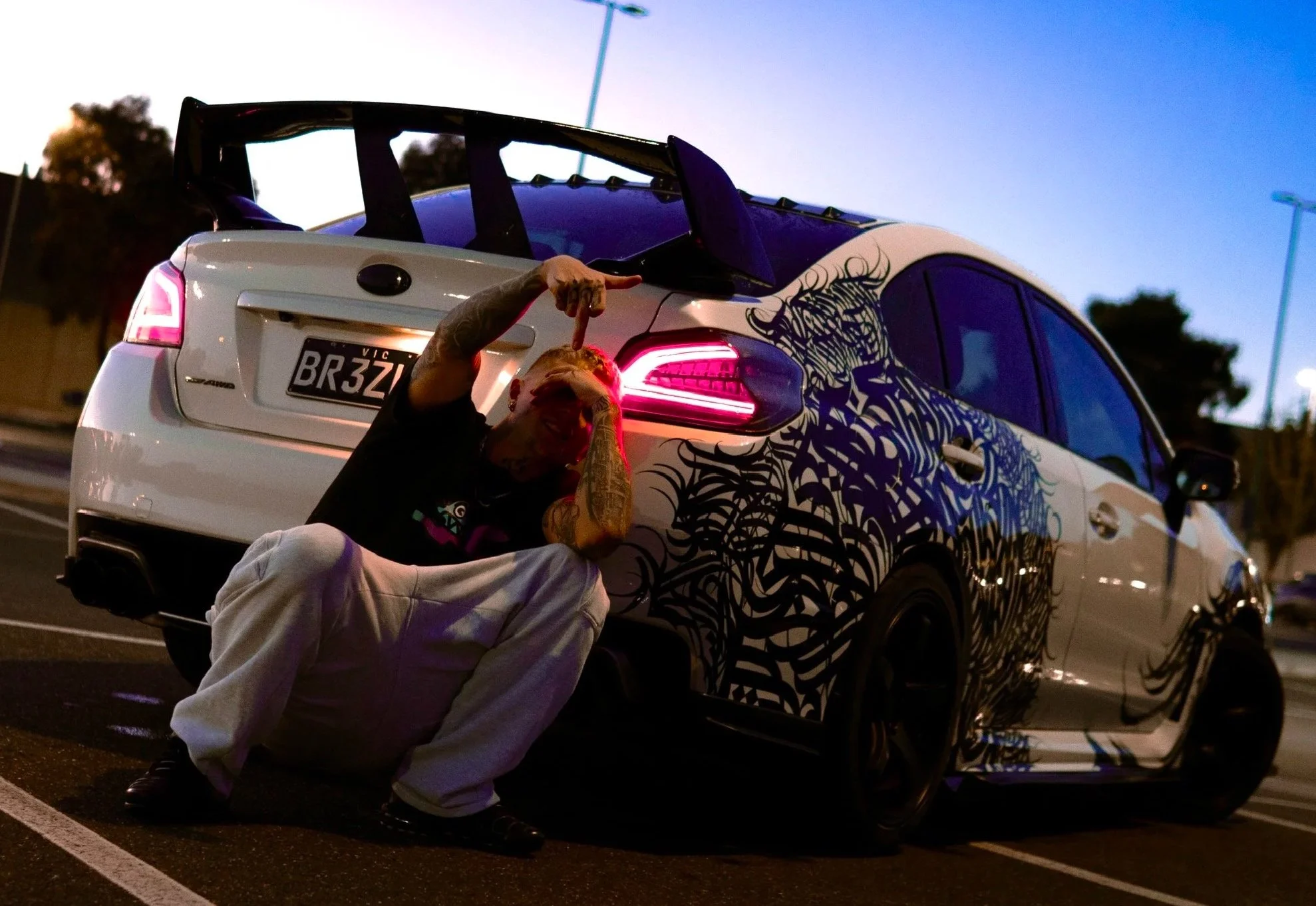 A person with tattoos kneeling next to a white car with black and blue artwork on the side, in a parking lot during dusk. The person appears to be pointing toward the camera and is smiling.