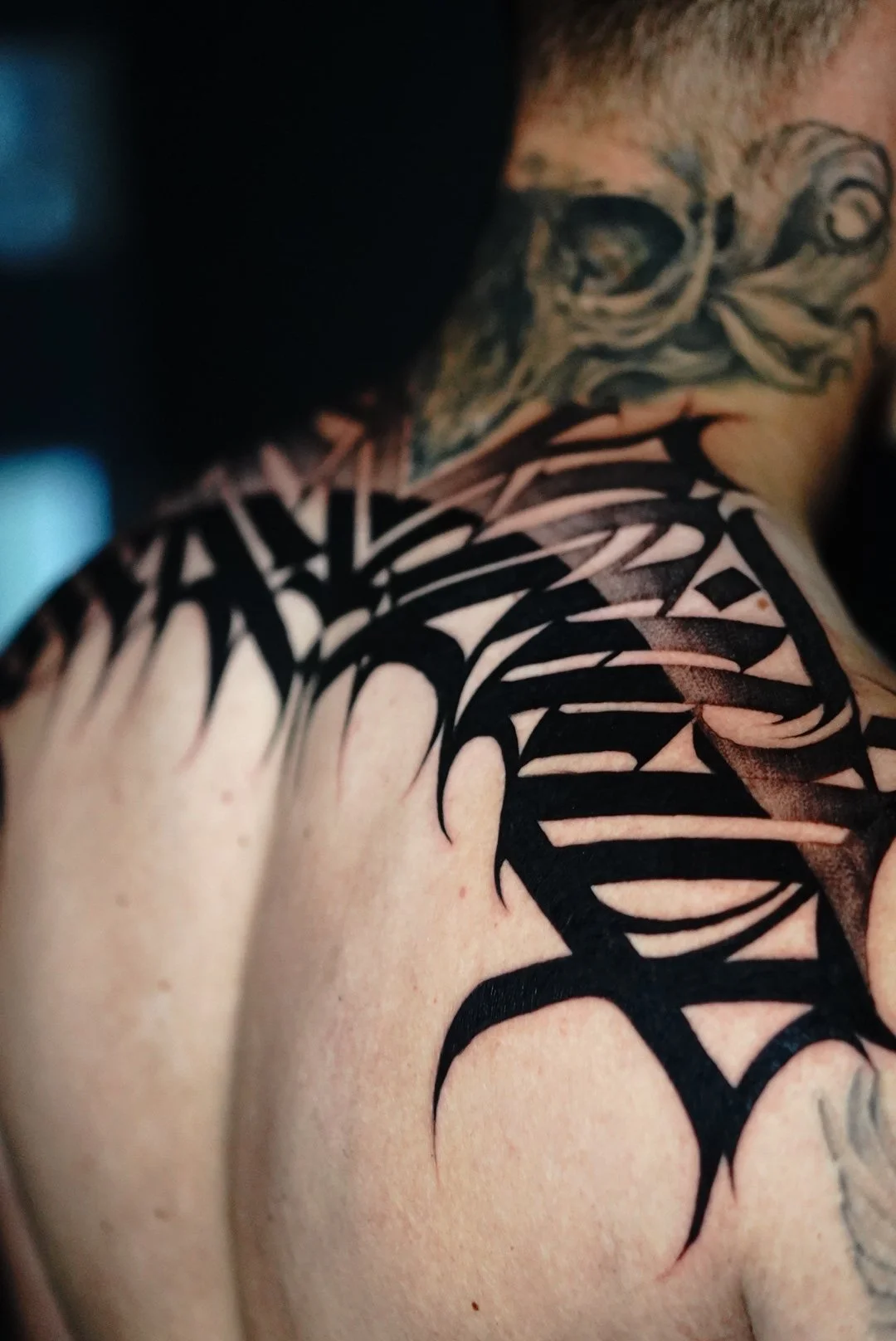 Close-up of a person's shoulder with black tribal-style tattoo and part of their face with another tattoo near the eye.
