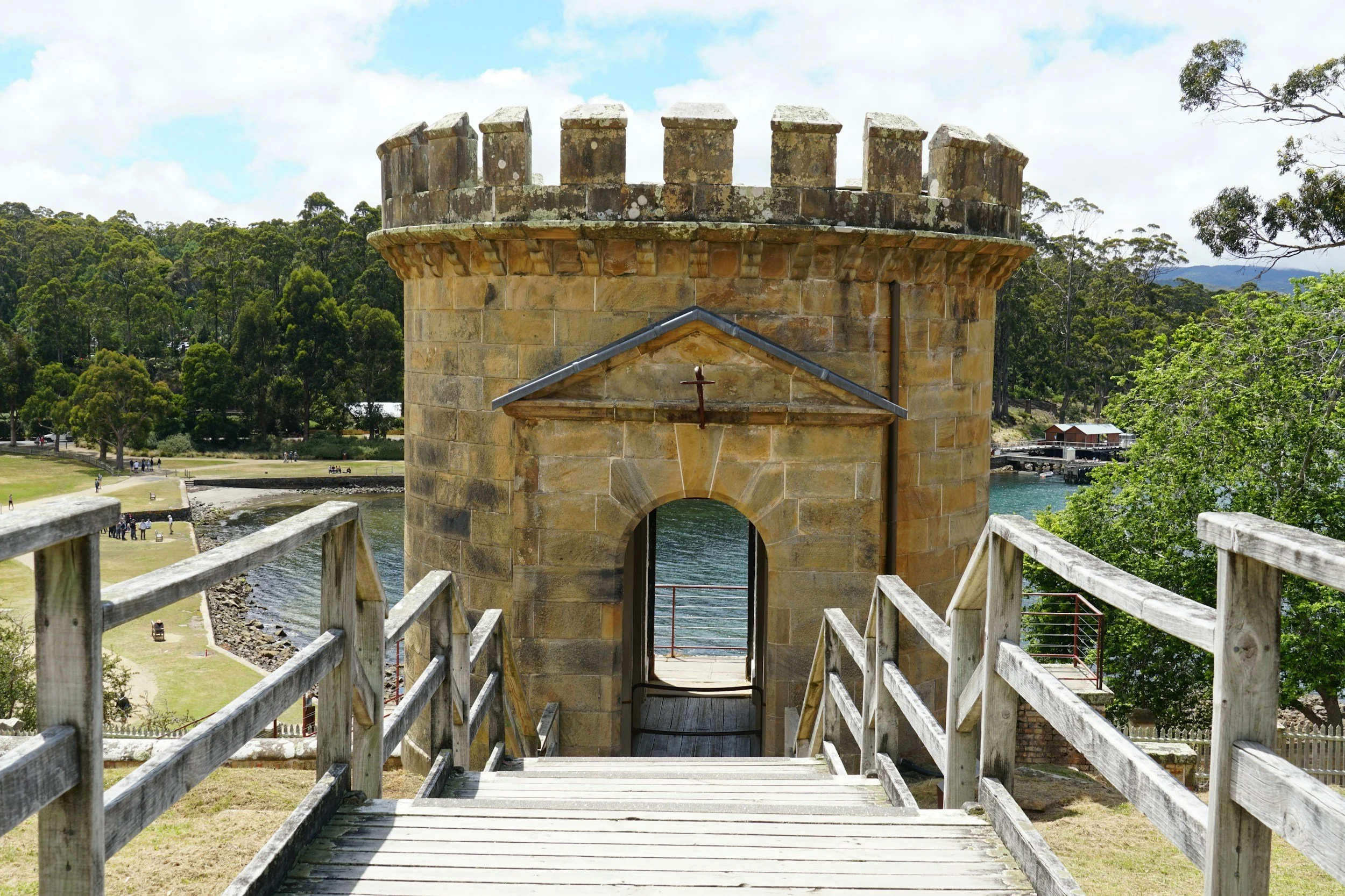 A stone tower with a crenellated top, viewed from a wooden walkway over water. Trees surround the area, and the sky is partly cloudy.