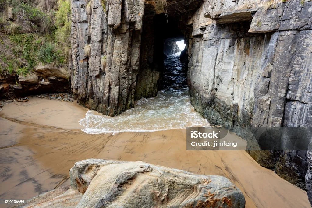 Rock formation with a natural archway, ocean waves hitting the sandy beach below, and water flowing through the arch into the sea, surrounded by cliffs.