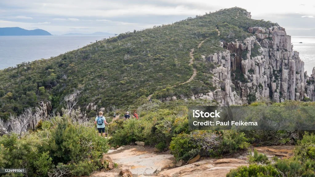 Hikers walking along a trail through a mountainous landscape with rocky cliffs and greenery, overlooking a body of water.
