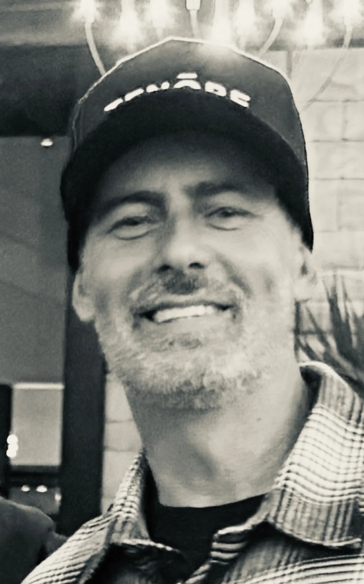 Close-up photo of a smiling man with short hair, beard, and mustache, wearing a baseball cap and a checkered jacket, in black and white.