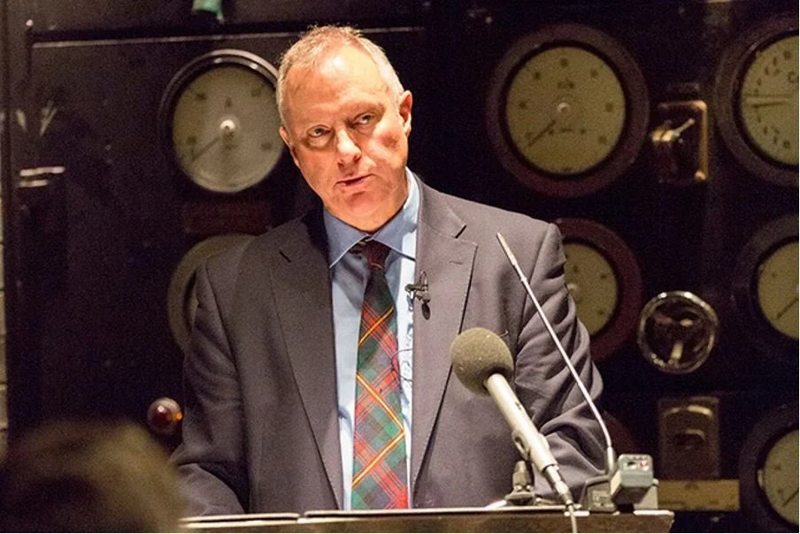 A man in a suit and tie speaking into a microphone at a press conference or public event, with large gauges on the wall behind him.