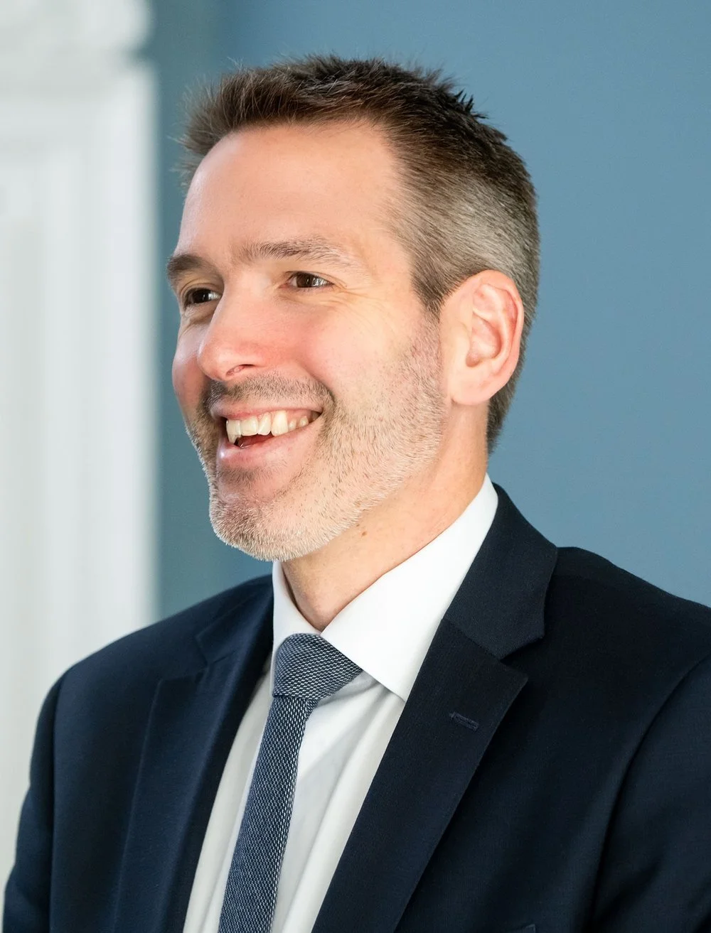 A smiling man with short, dark hair and a beard, wearing a dark suit and a white shirt with a patterned tie, standing indoors against a blue wall.