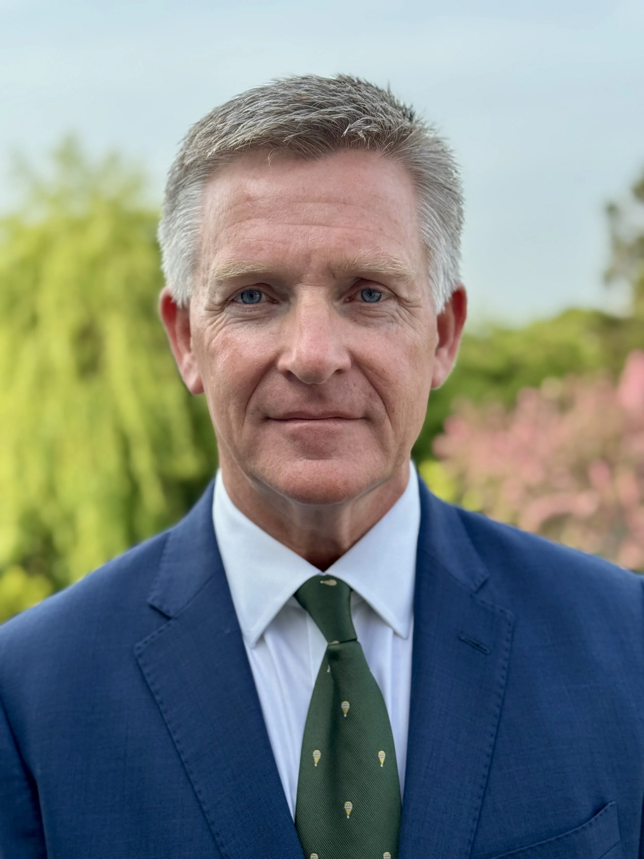 A middle-aged man with salt-and-pepper hair, wearing a dark blue suit, white shirt, and a green tie with ice cream cone patterns, standing outdoors with blurred greenery in the background.
