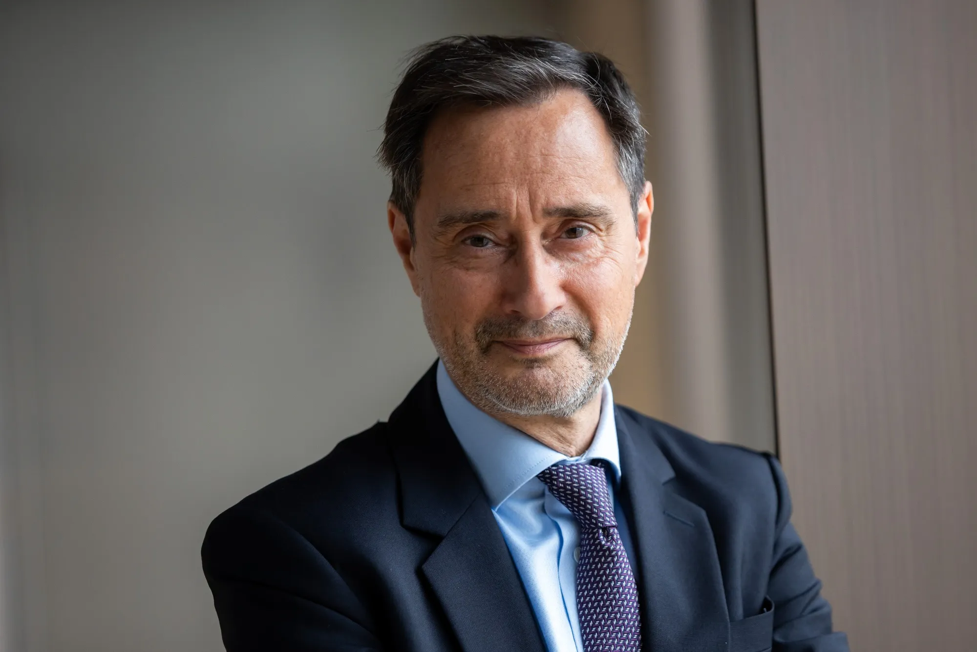 A middle-aged man with dark hair, grey streaks, and light skin wearing a dark suit, light blue shirt, and purple tie, standing near a window indoors.