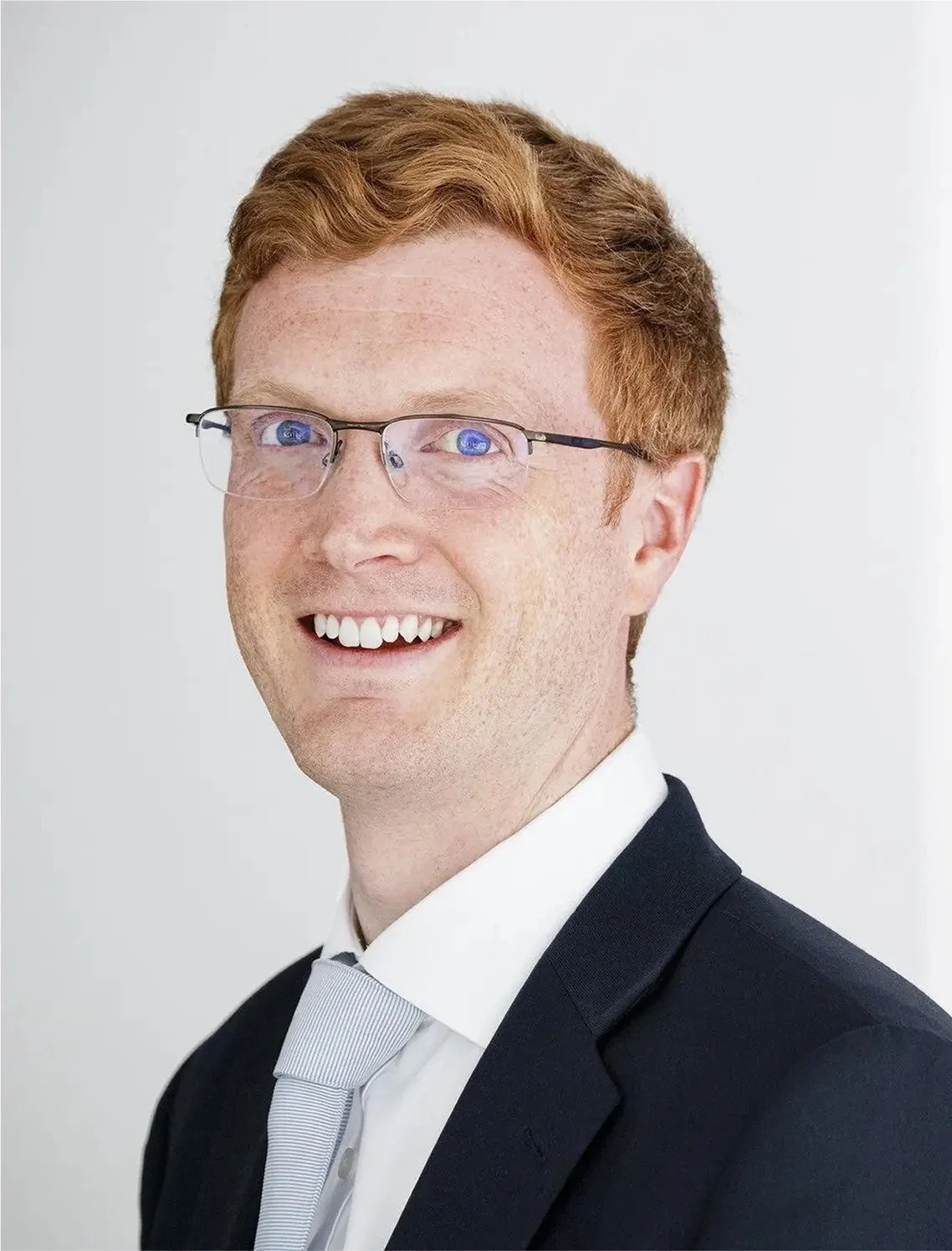 A smiling man with red hair, glasses, wearing a black suit, white shirt, and light-colored tie, against a light background.