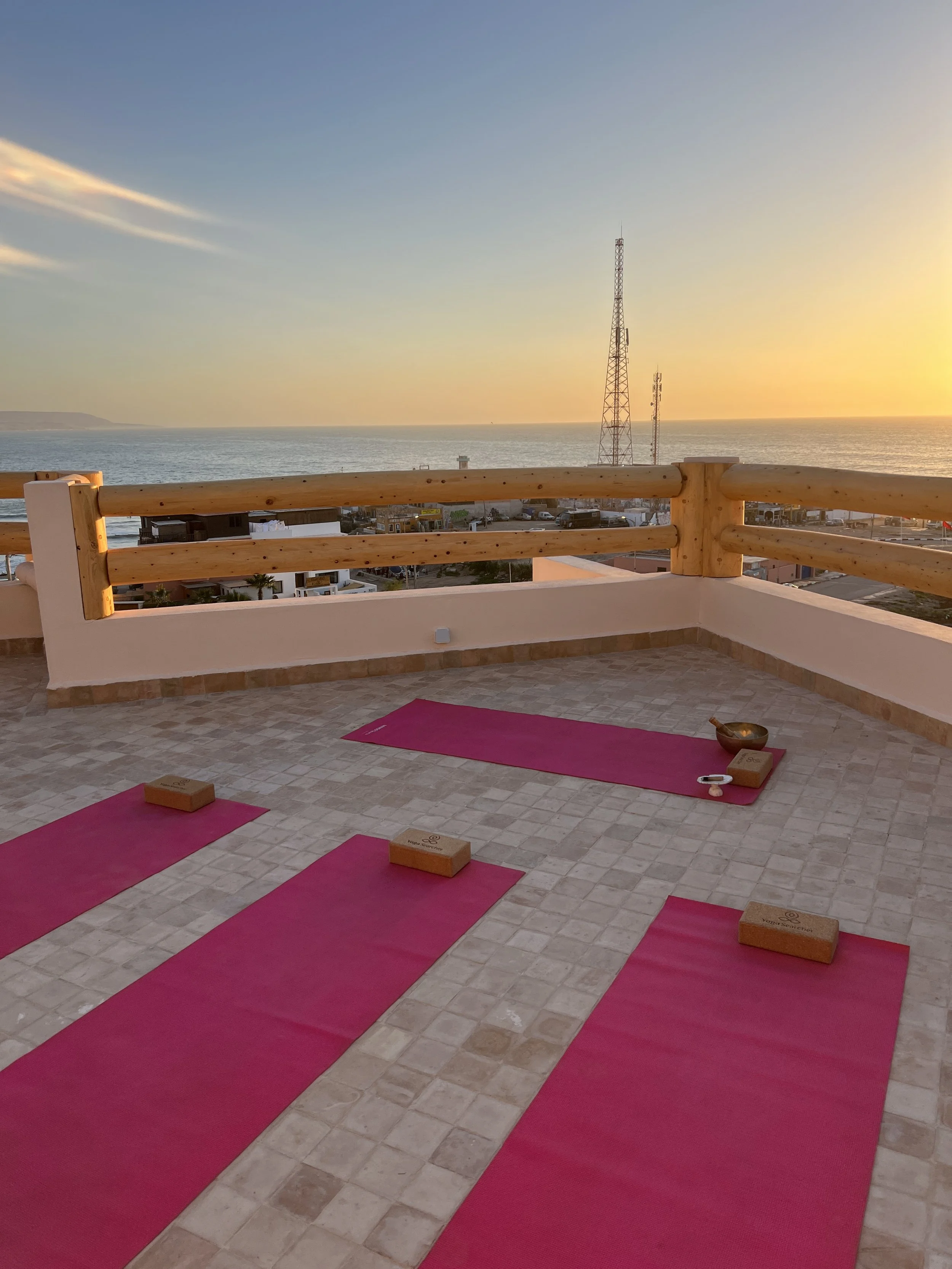 Un balcon avec des tapis de yoga roses, des briques en bois pour yoga, un bol tibétain, un bâton de massage en bois, et une vue sur la mer au coucher du soleil.
