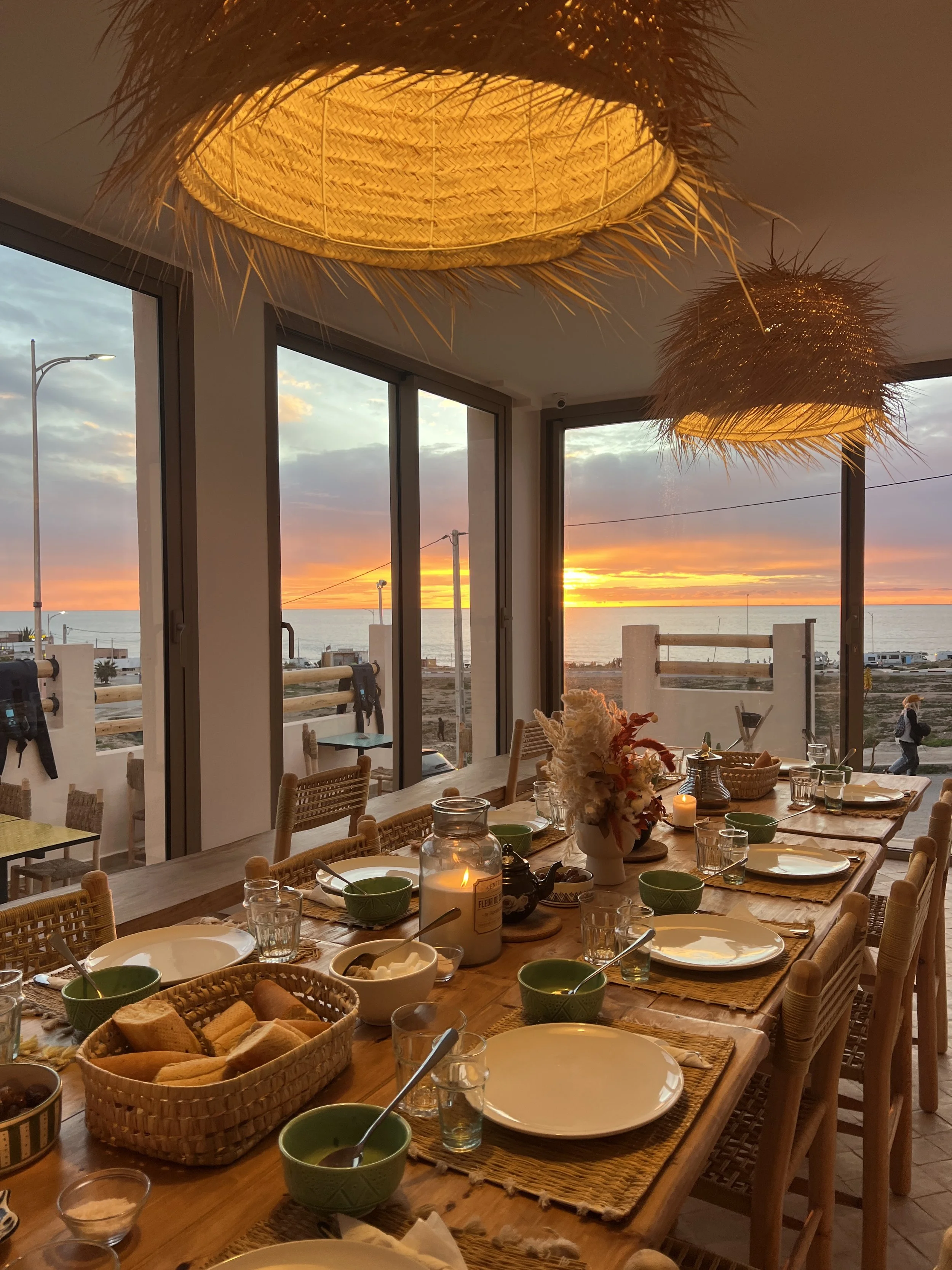 Une salle à manger avec une grande table en bois décorée avec des bougies, des fleurs et des assiettes, face à une baie vitrée donnant sur le coucher de soleil sur la mer, éclairée par des lampes en osier suspendues au plafond.