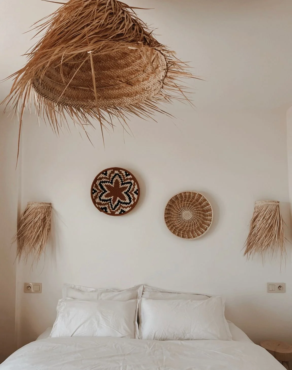Un mur blanc décoré de trois paniers en osier suspendus, un au centre avec un motif floral noir et blanc, et deux autres avec des motifs géométriques, avec deux appliques en paille en forme de demi-cercle de chaque côté, au-dessus d'un lit avec des draps blancs.