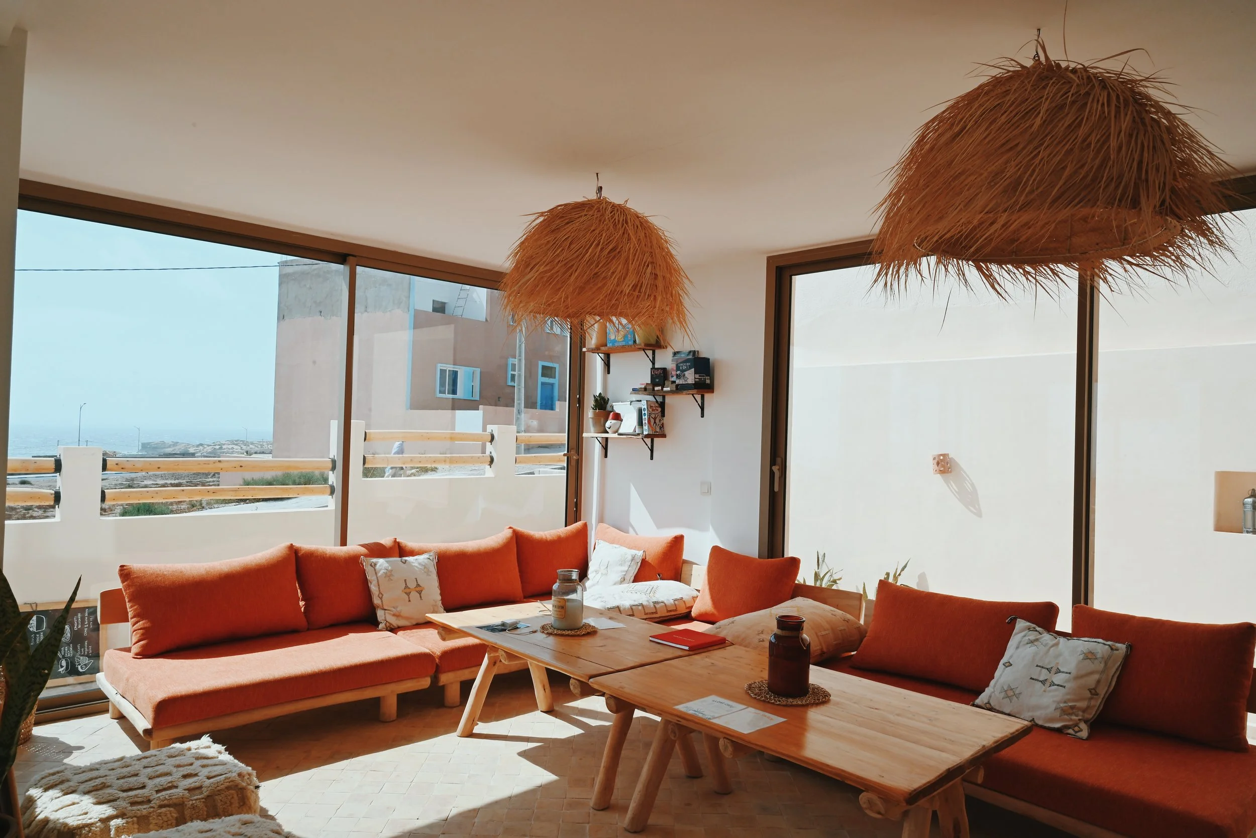 Intérieur d'un salon avec un canapé en tissu orange, une table en bois, un canapé en bois, des coussins avec motifs, des lampes en matériaux naturels, de grandes fenêtres laissant entrer la lumière naturelle et offrant une vue sur un bâtiment en extérieur.